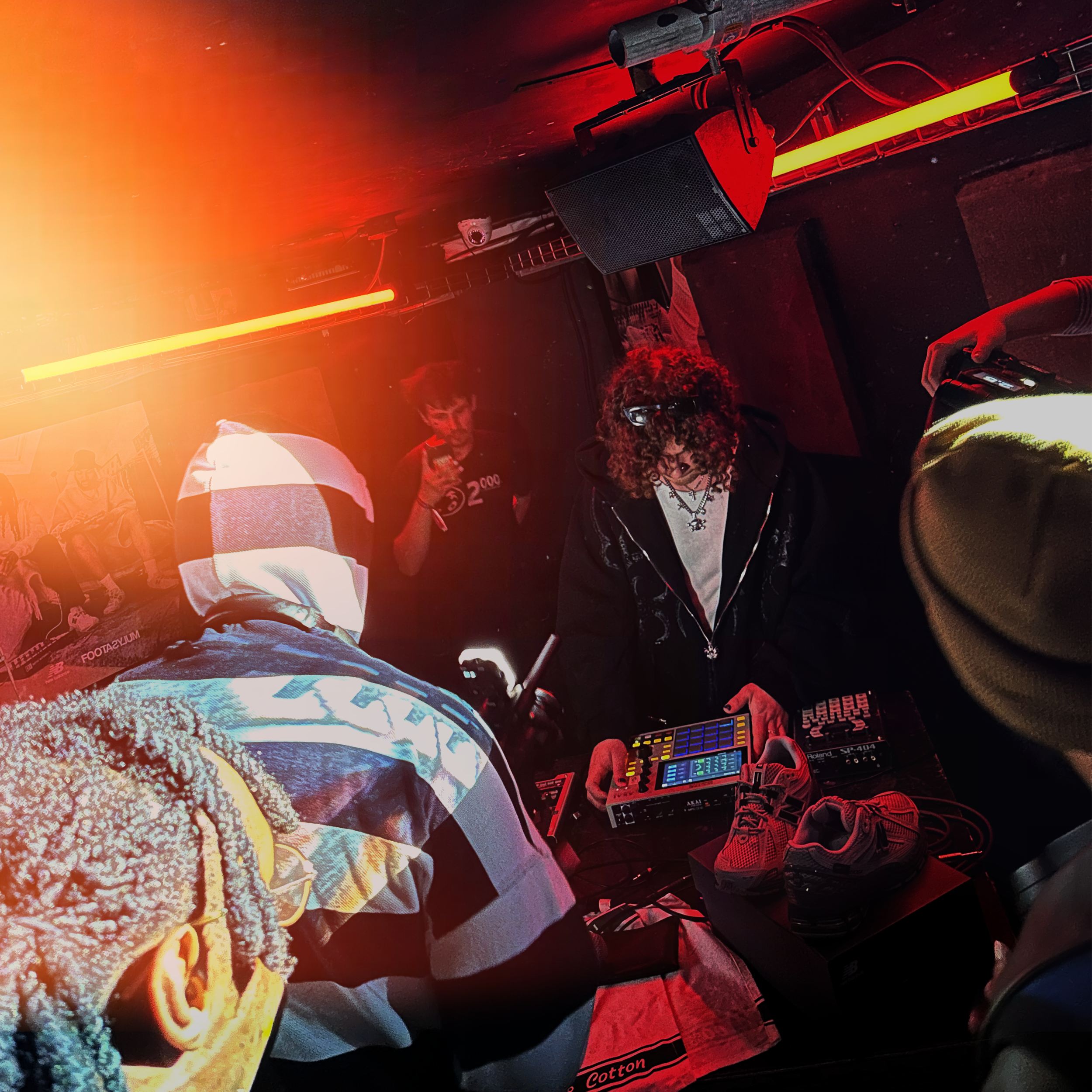 The image shows a DJ setup with a mixer and electronic equipment, illuminated by red lighting. There are shoes on the floor, and a person is operating the equipment wearing a black hoodie, with their hand interacting with the device. The environment 