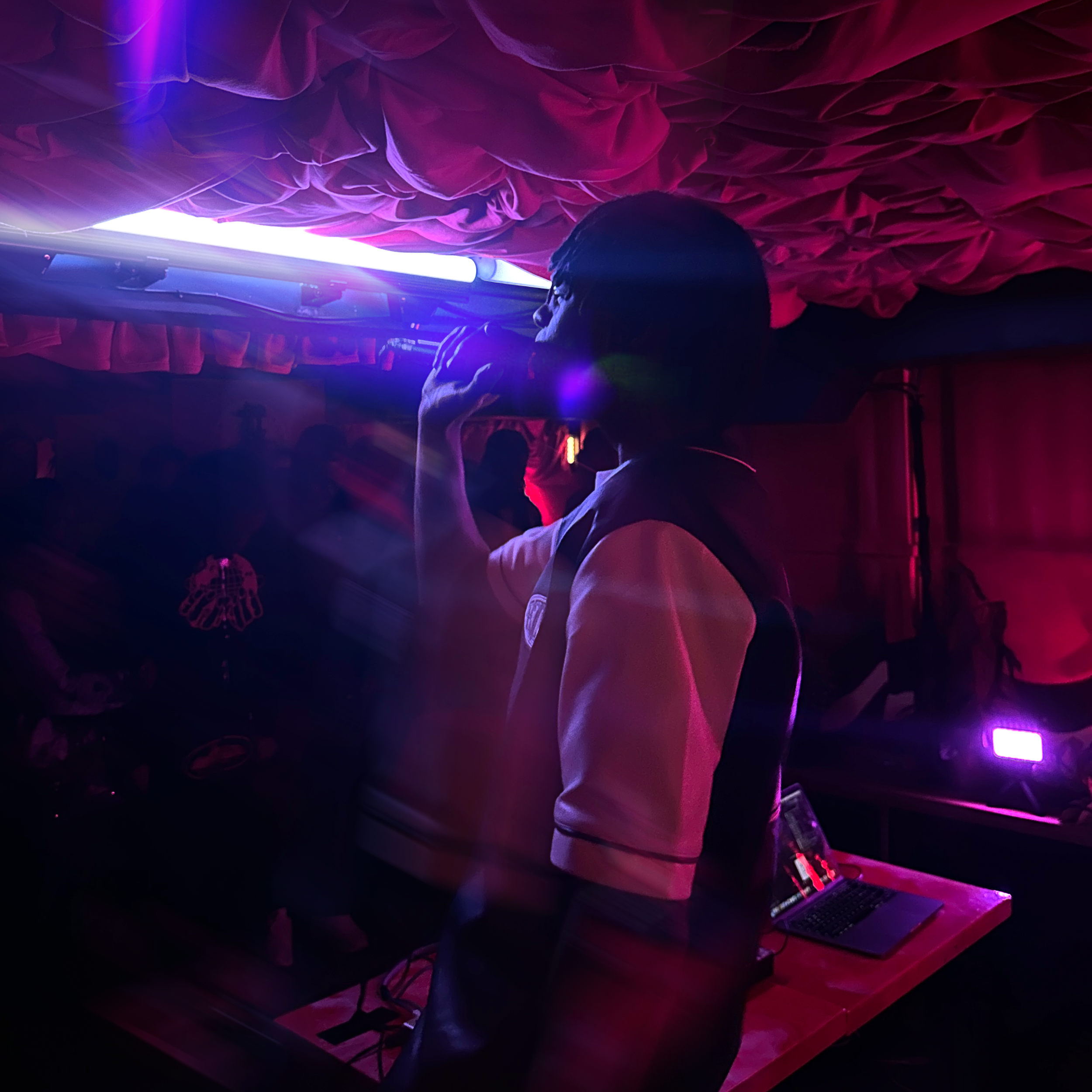 Person using a laptop at a DJ booth with colorful pink and purple lighting and a fabric backdrop.