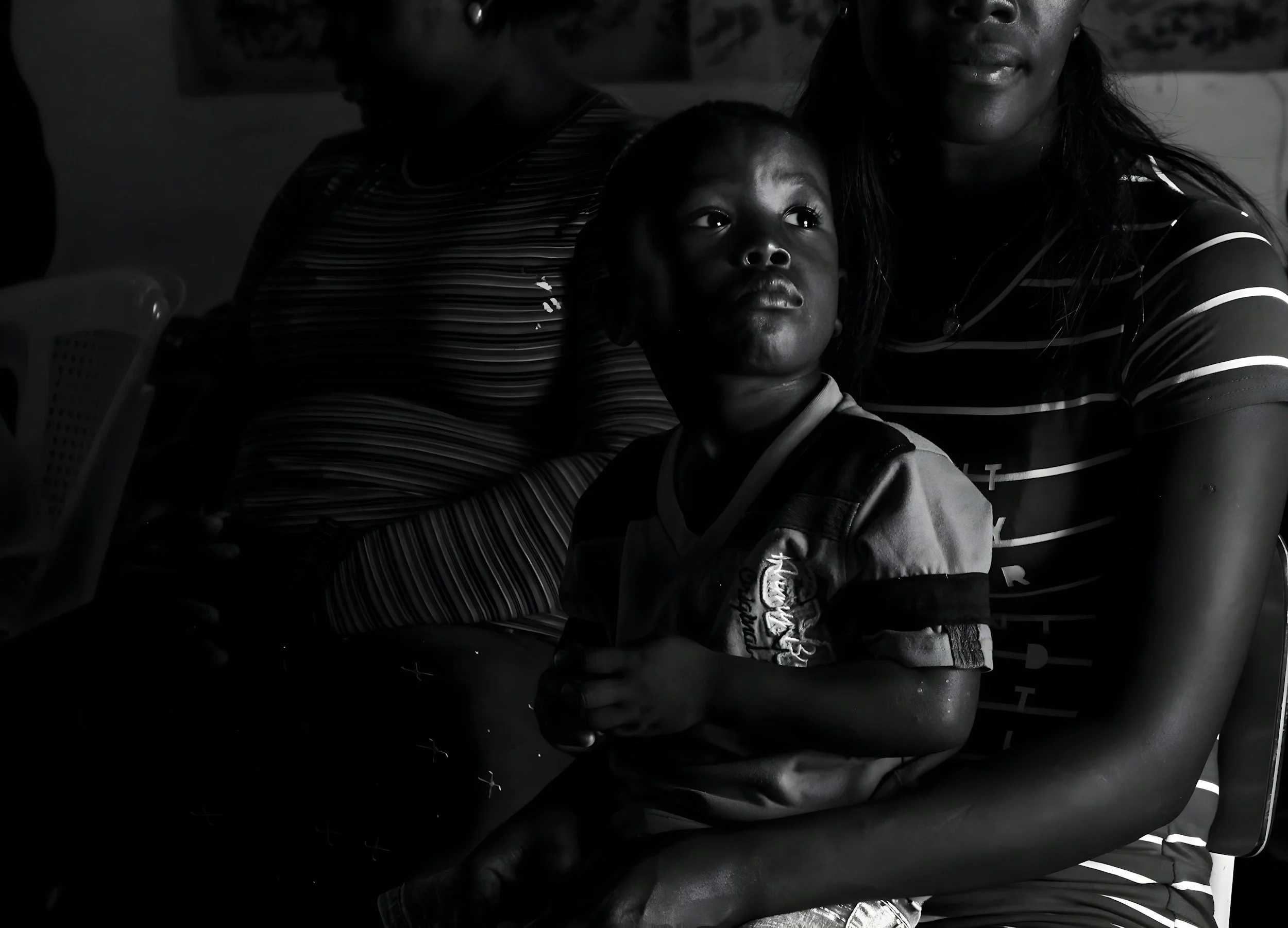 A black and white photograph of a pensive young boy sitting on his mother's lap in front of another woman, reflecting ancestral family dynamics.