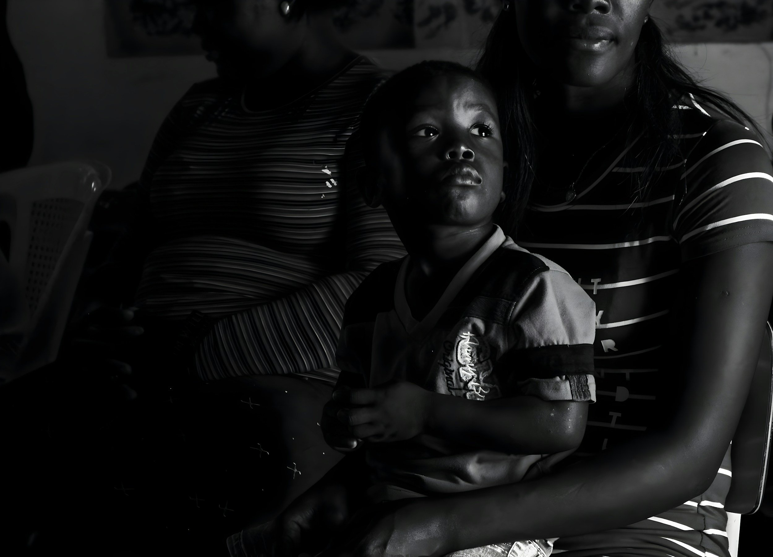 A black and white photograph of a pensive young boy sitting on his mother's lap in front of another woman, reflecting ancestral family dynamics.