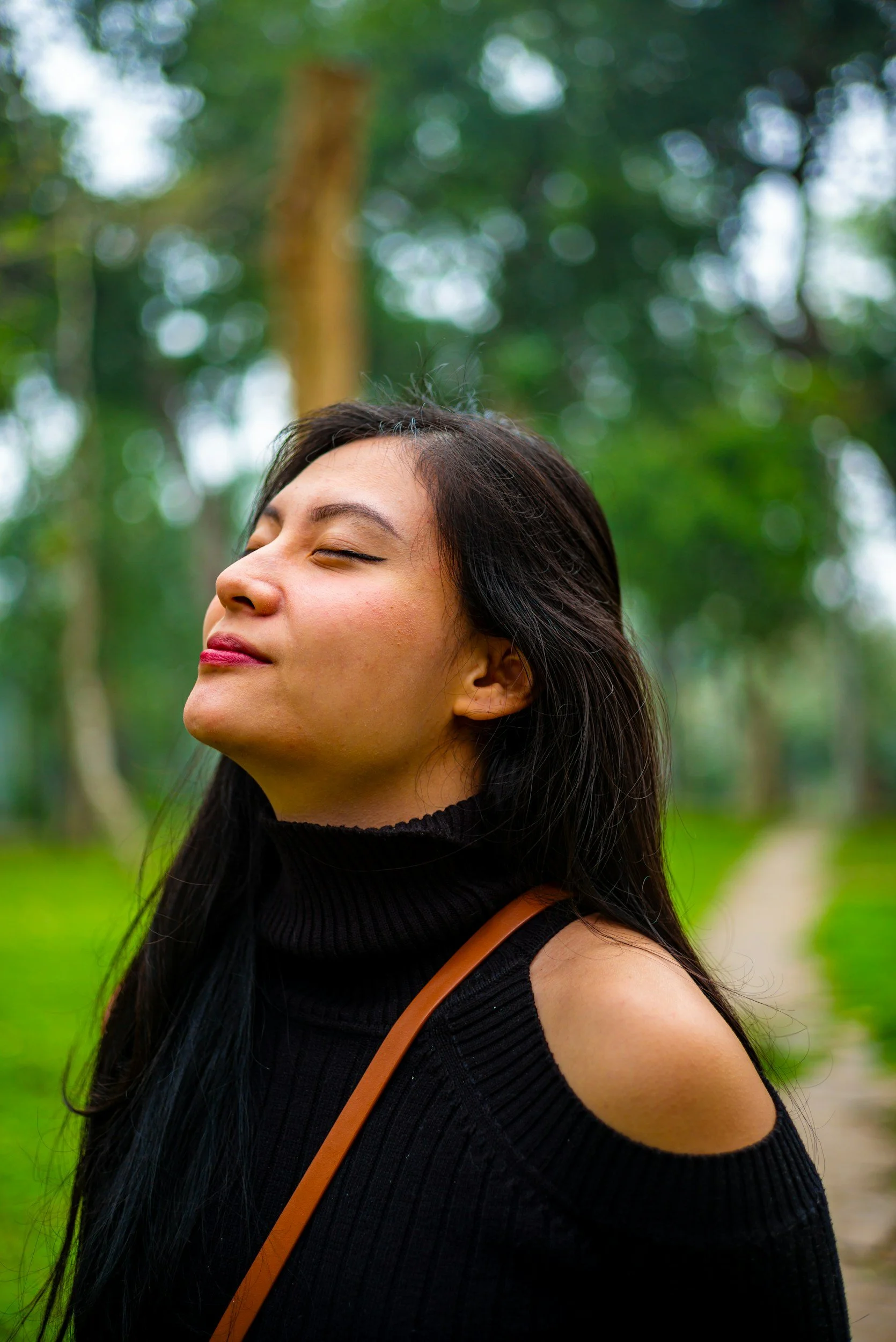 A woman in a lush green forest looking up at the sky with her eyes closed and a soft smile, practicing mindful grounding.