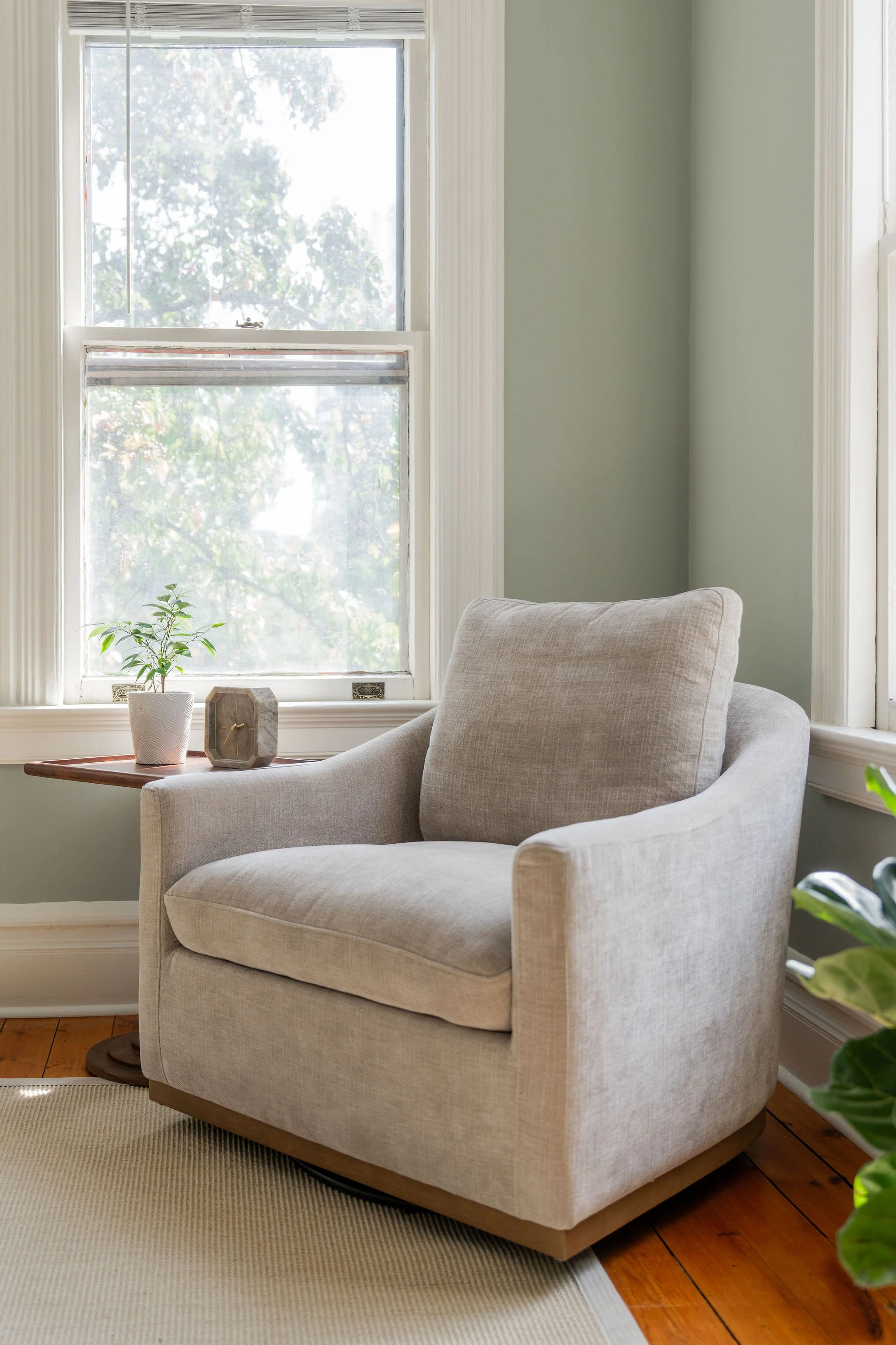 Gray club chair in front of two windows with side table with clock and plant next to it