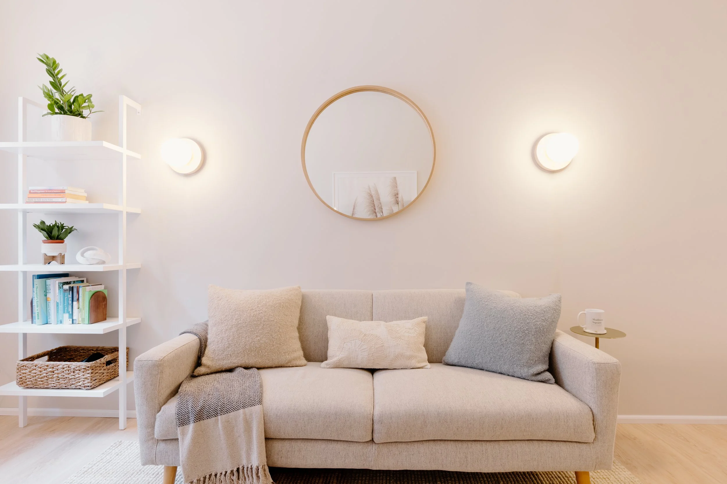 A cozy living room with a beige sofa adorned with throw pillows, a blanket, and a small side table holding a mug. A round mirror hangs on the wall above the sofa, flanked by two wall-mounted lights. To the left, a white shelving unit displays books and decorative plants.