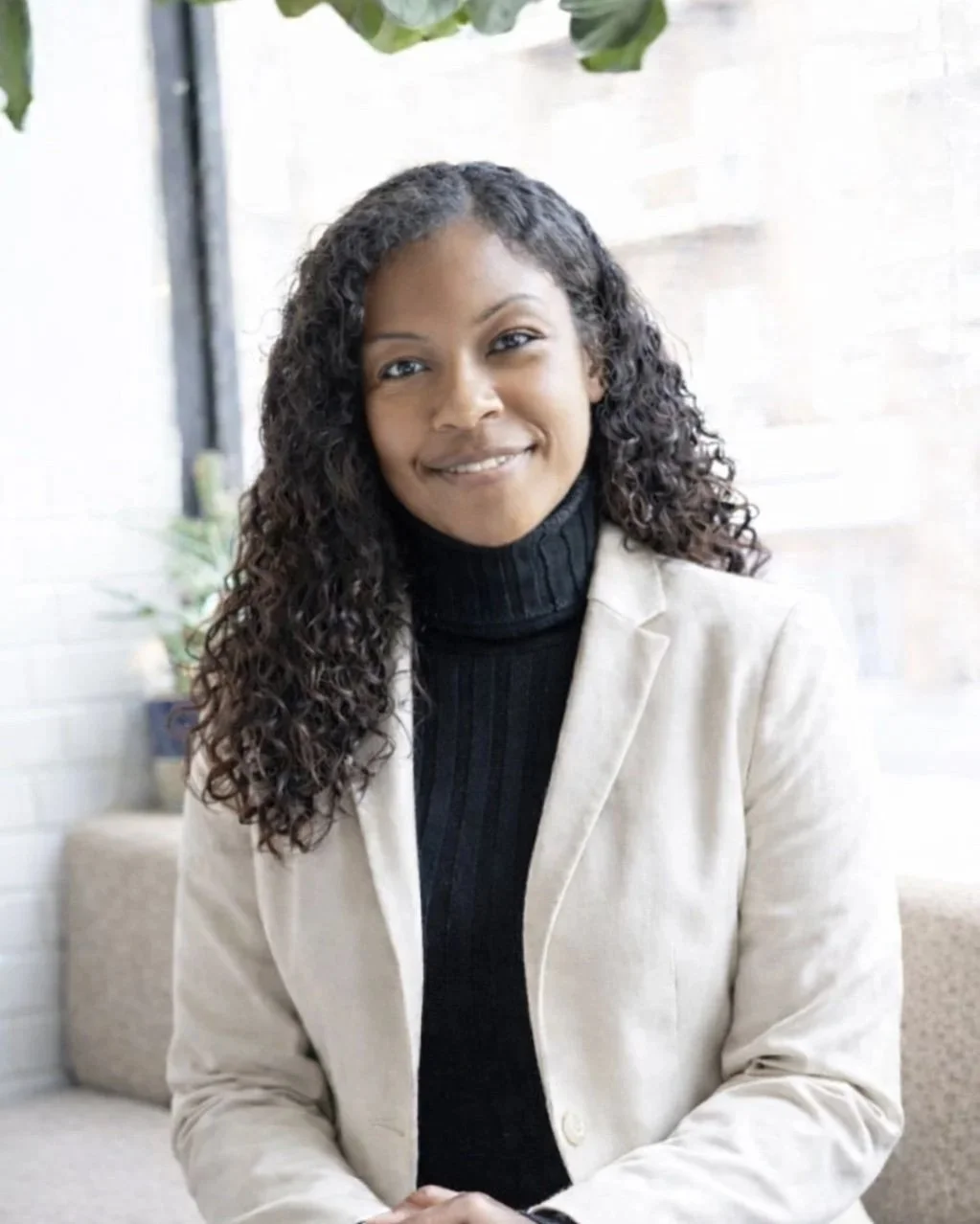 A woman with long dark hair smiling at the camera, wearing a black shirt and a cream blazer