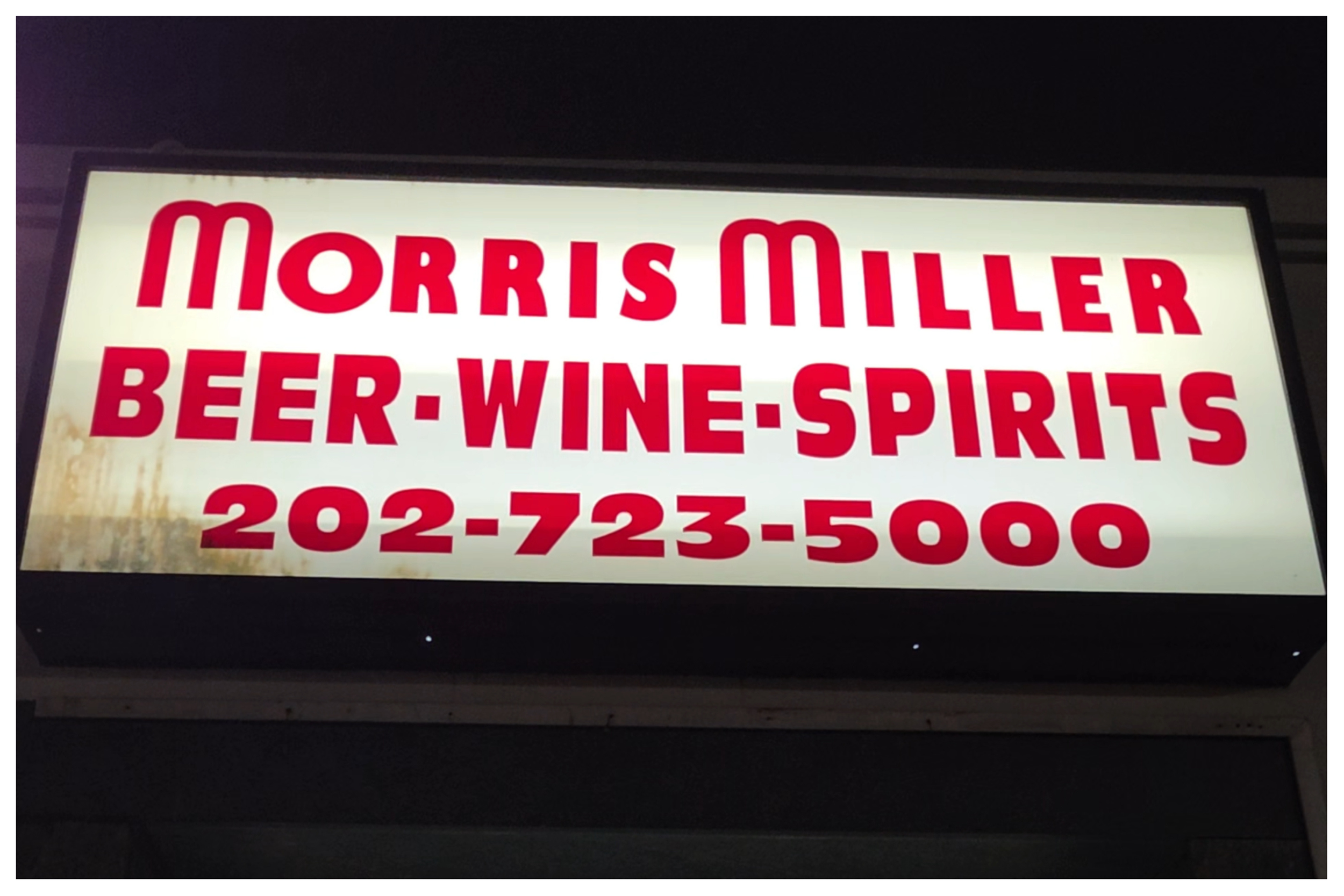 A brightly lit sign displaying the name 'Morris Miller' in large red letters, with additional text for beer, wine, and spirits, and a contact phone number.