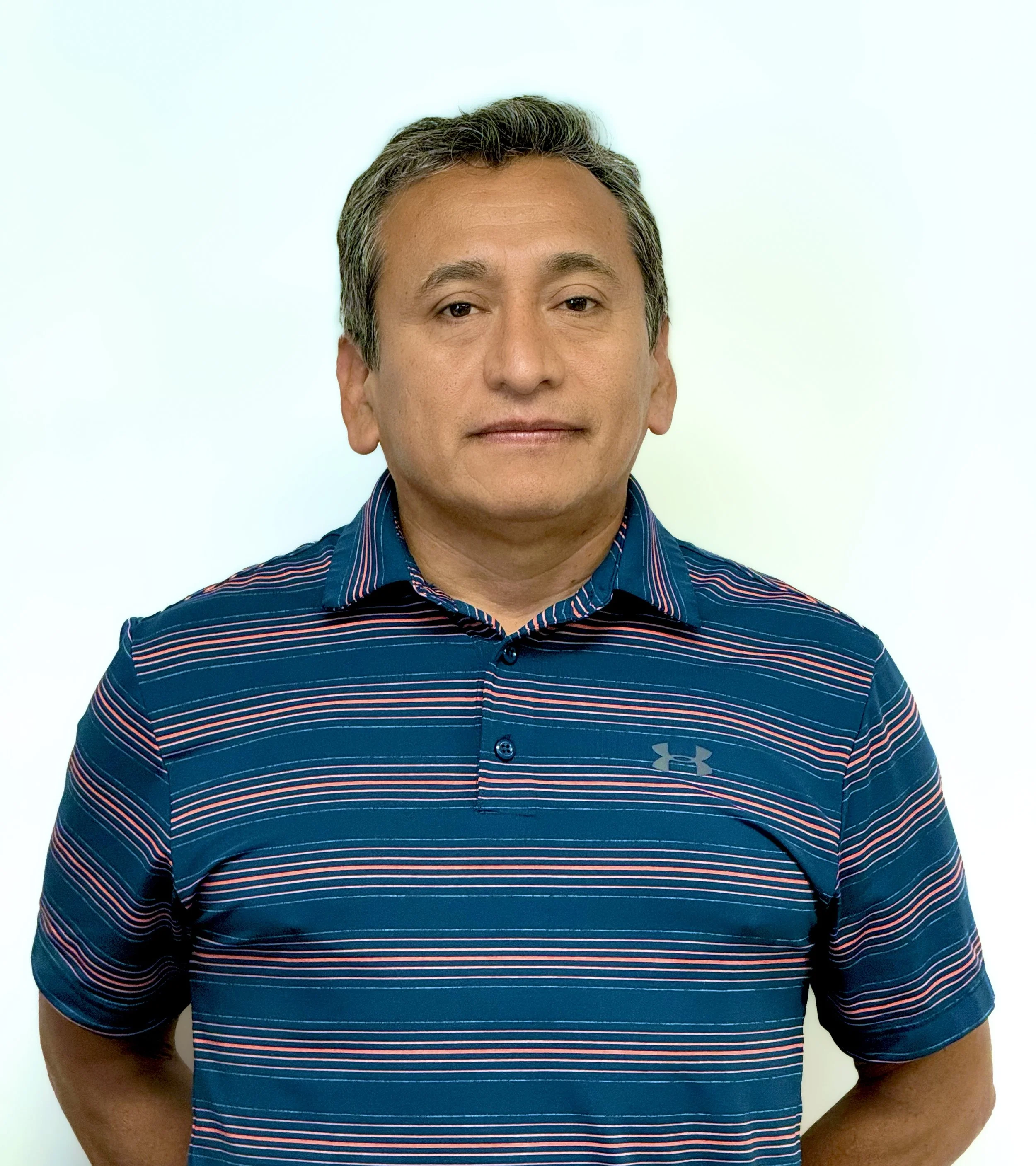 A middle-aged man with short, dark, graying hair wearing a blue, red, and white striped Under Armour polo shirt standing against a plain white background.
