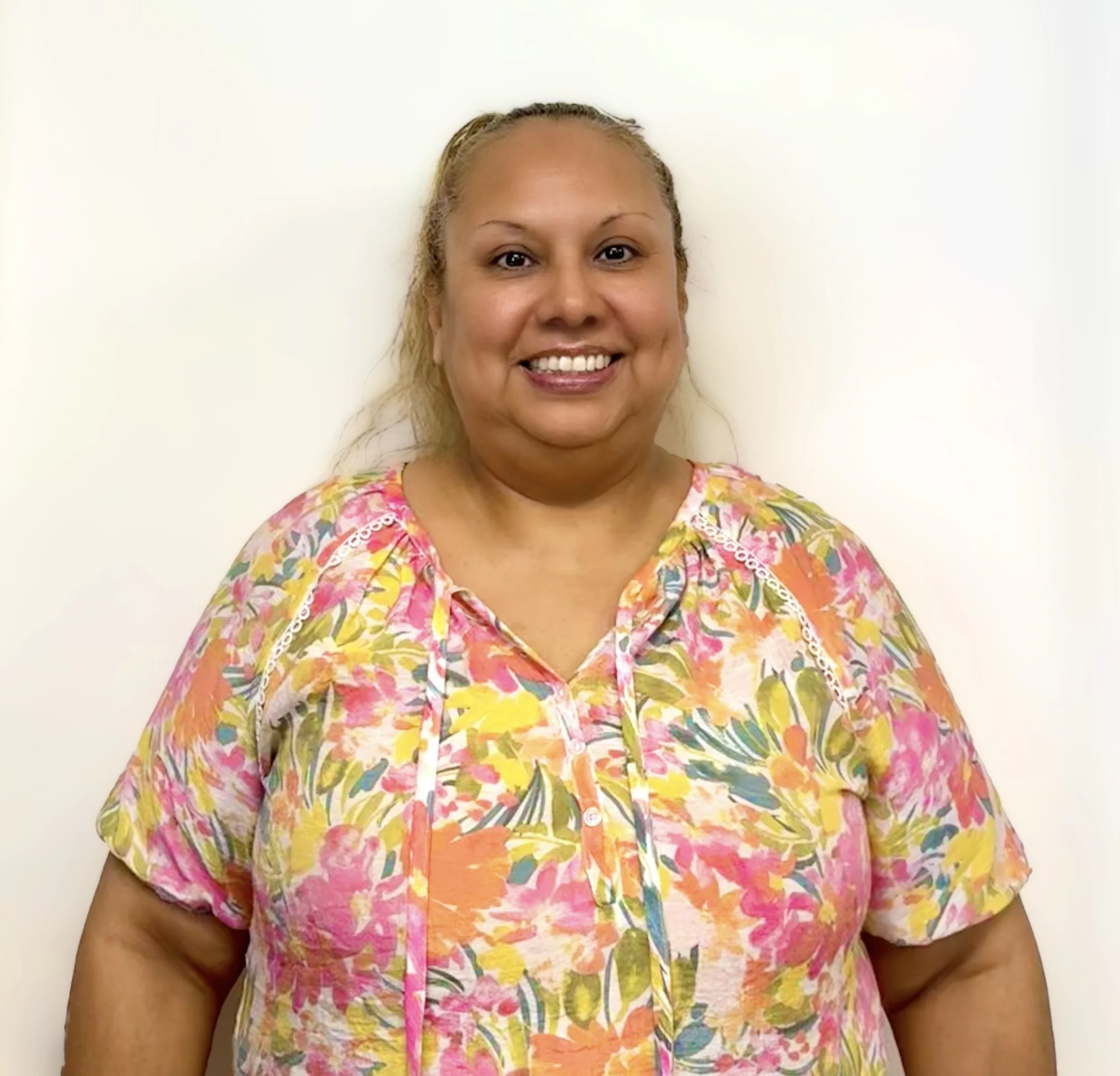 Smiling woman in colorful floral shirt standing against a plain white wall.