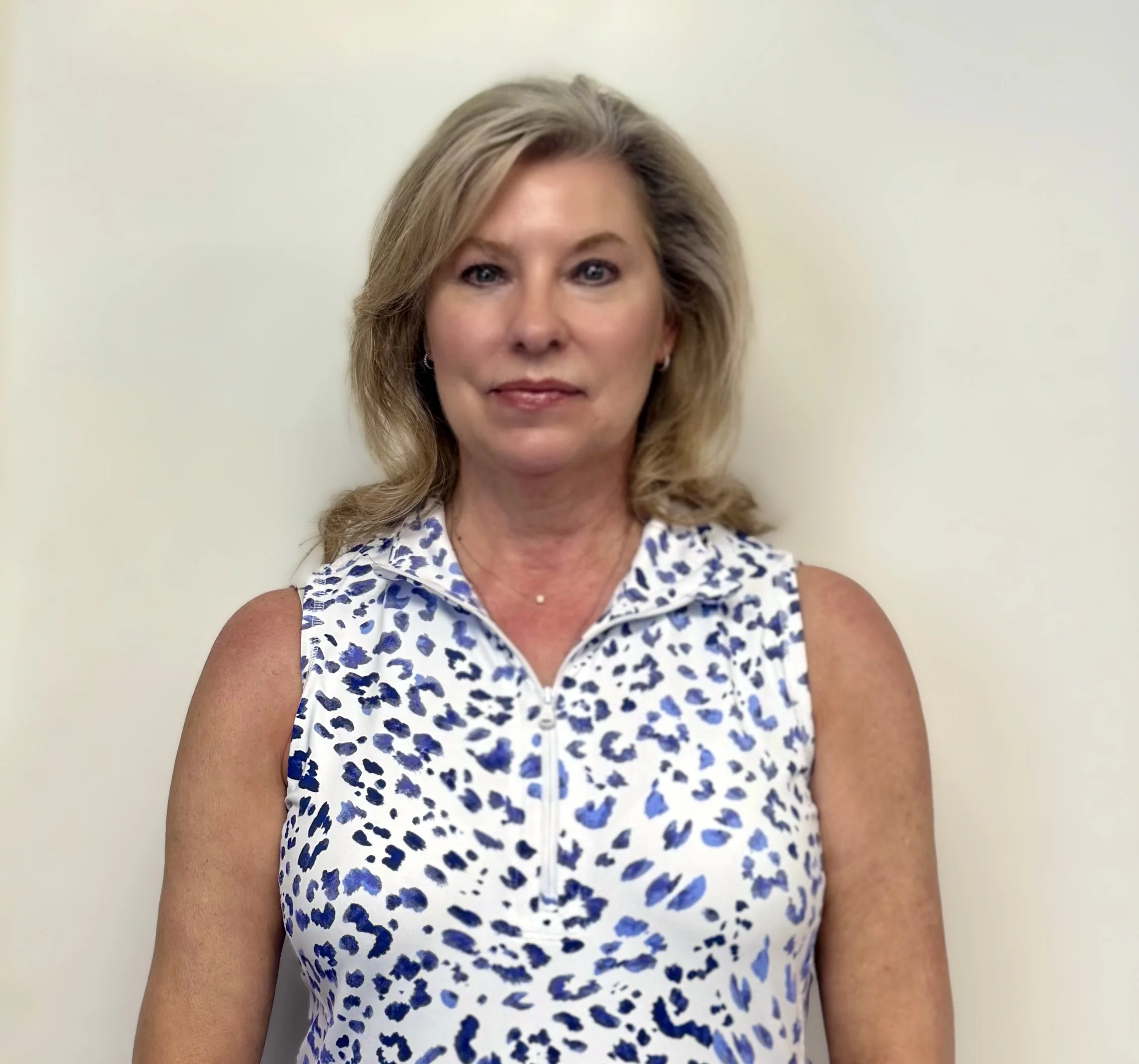A woman with blonde hair wearing a sleeveless white and blue animal print top, standing against a plain off-white wall.