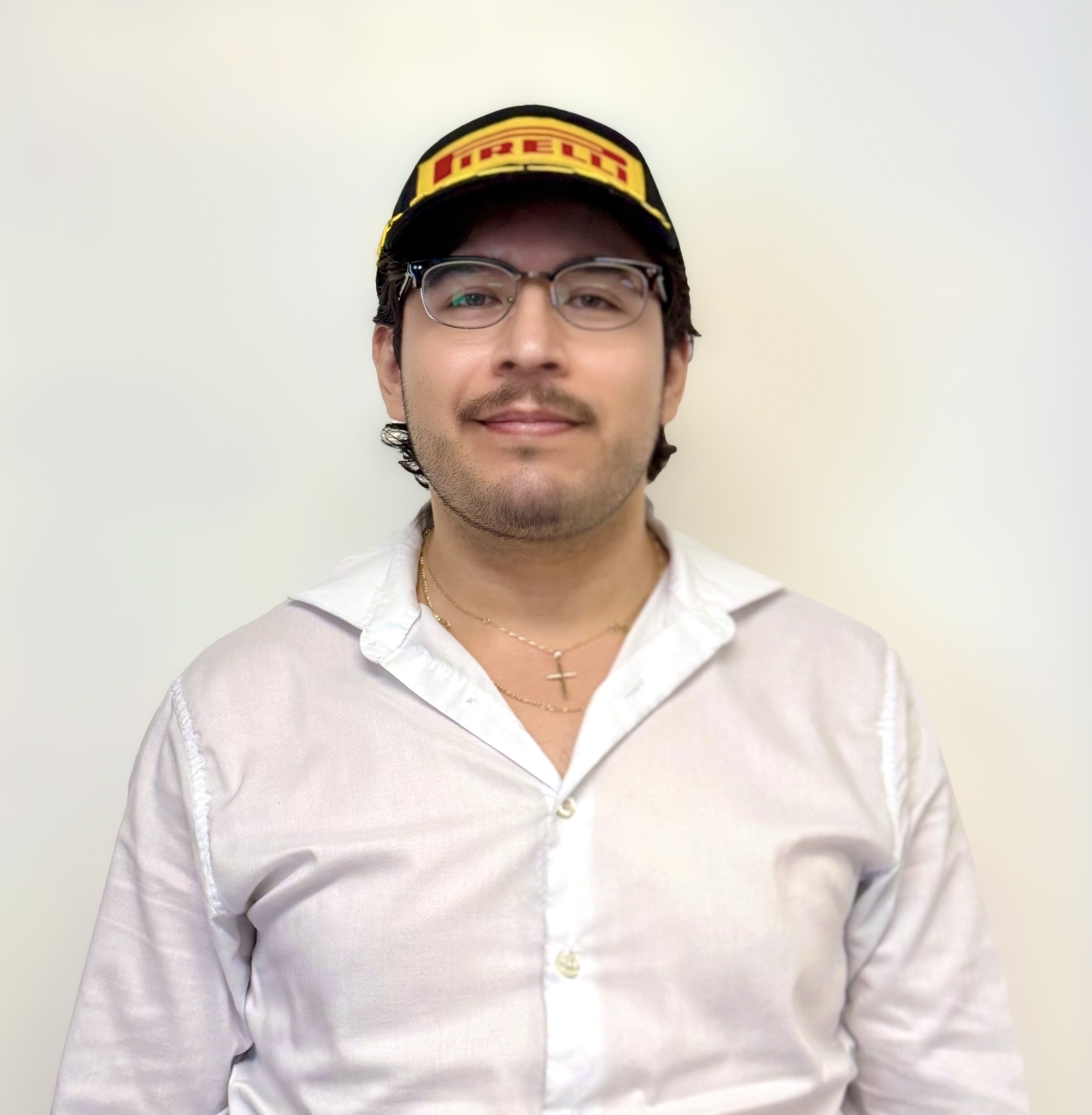 A man wearing glasses, a white shirt, a Pirelli cap, and a cross necklace standing against a plain wall.