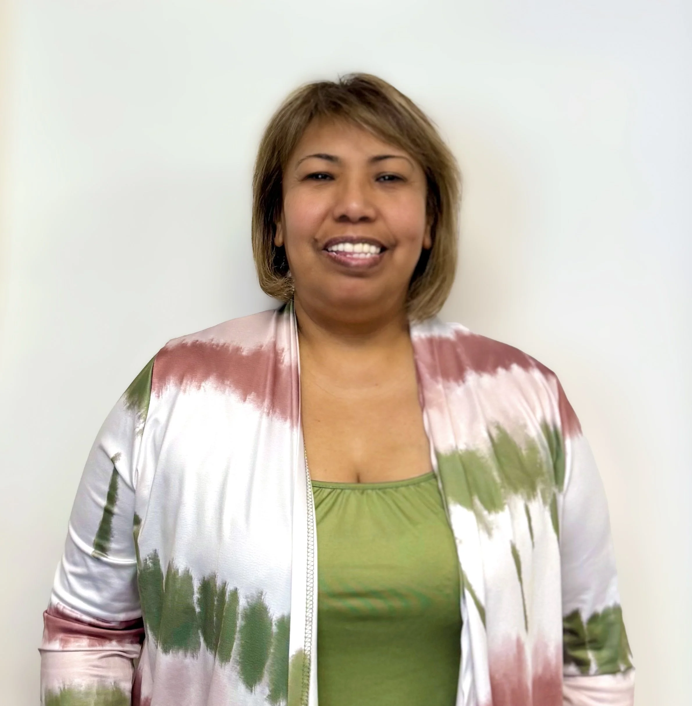 Woman with short hair smiling, wearing a green top and a multicolored satin jacket, standing against a plain white wall.
