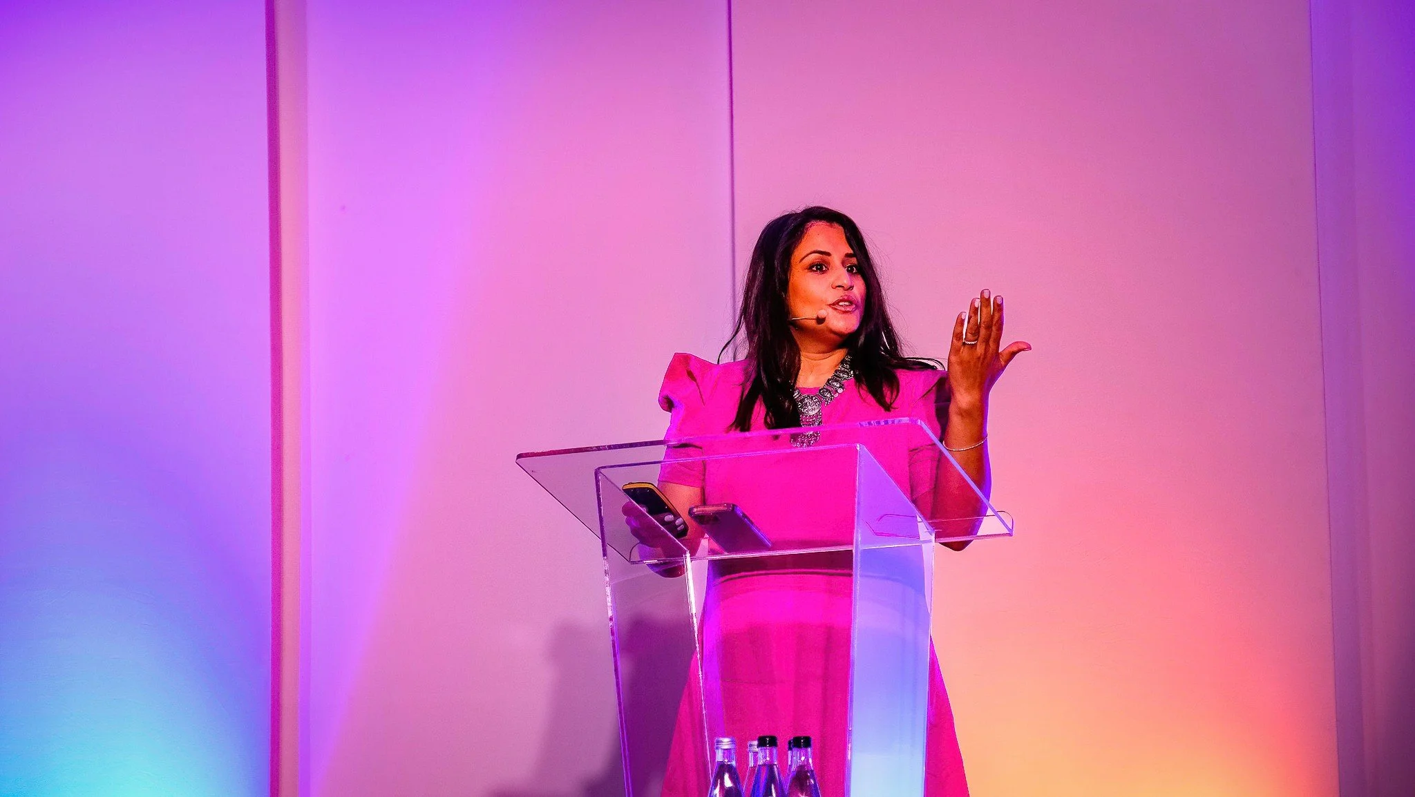 A woman in a pink dress speaking at a podium with a microphone and holding a phone, with a colorful background.
