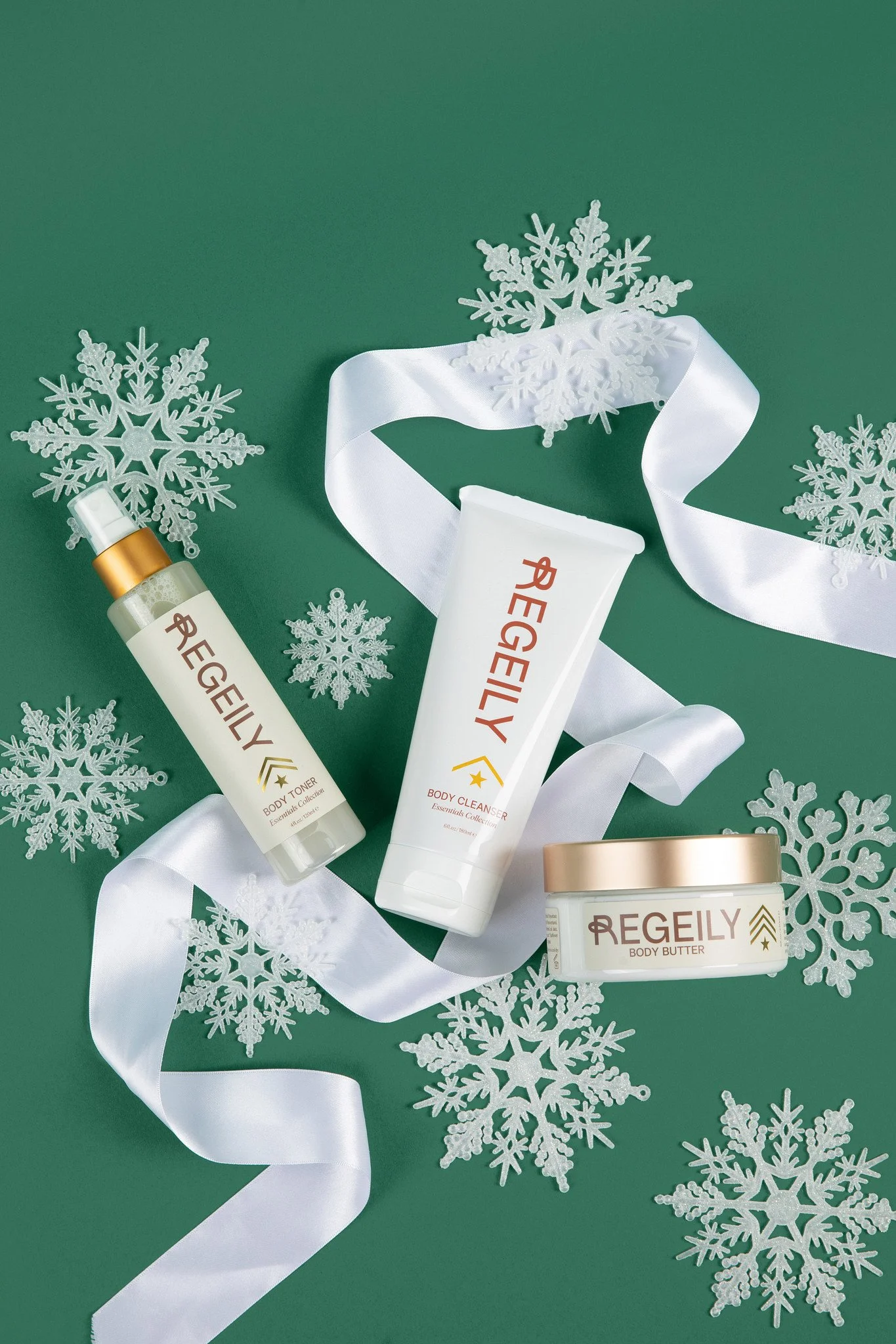 Regeily Body Cleanser, Body Toner, and Body Butter arranged with white ribbon and snowflakes on a green holiday background.