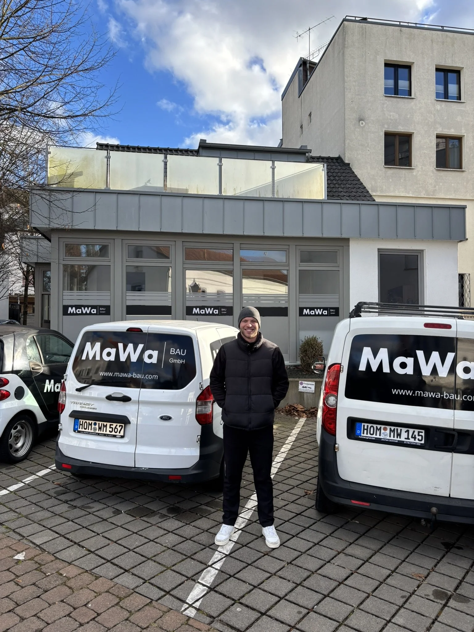 Ein Mann steht vor zwei weißen Firmenfahrzeugen mit MaWa Bau GmbH auf der Heckscheibe, im Hintergrund ein Gebäude mit mehreren Fenstern und blauen Himmel mit Wolken.