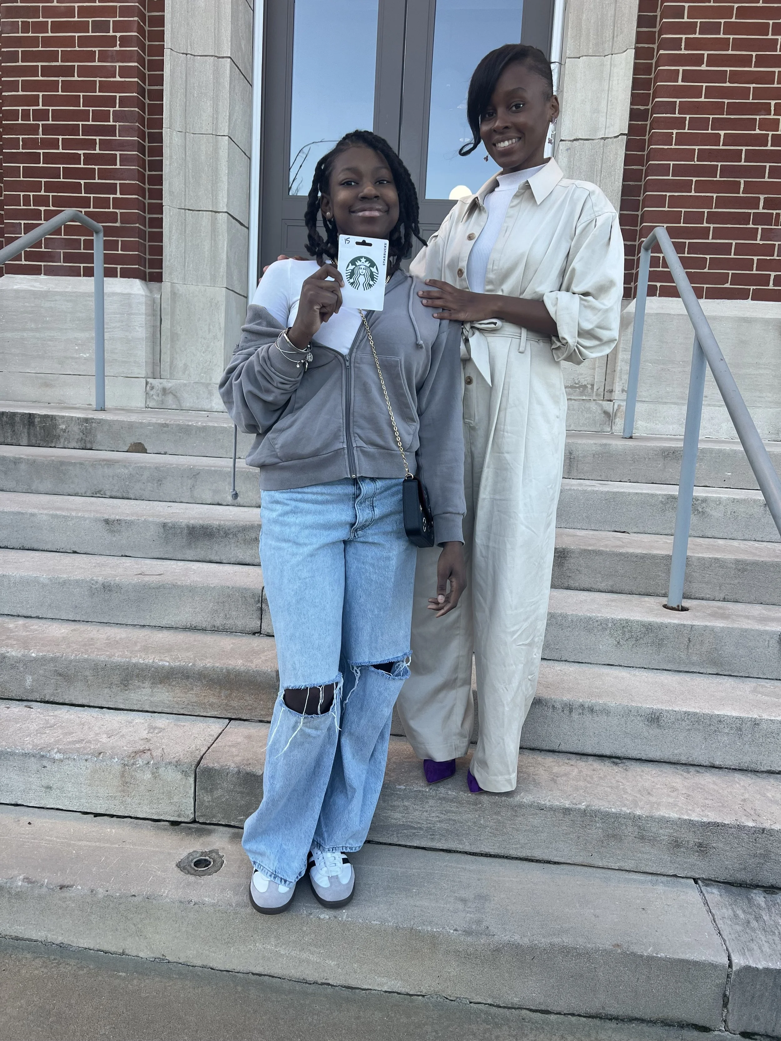 Two women standing on steps outside a building, smiling at the camera. One woman holds a Starbucks gift card, and the other has her hand on the first woman's shoulder.