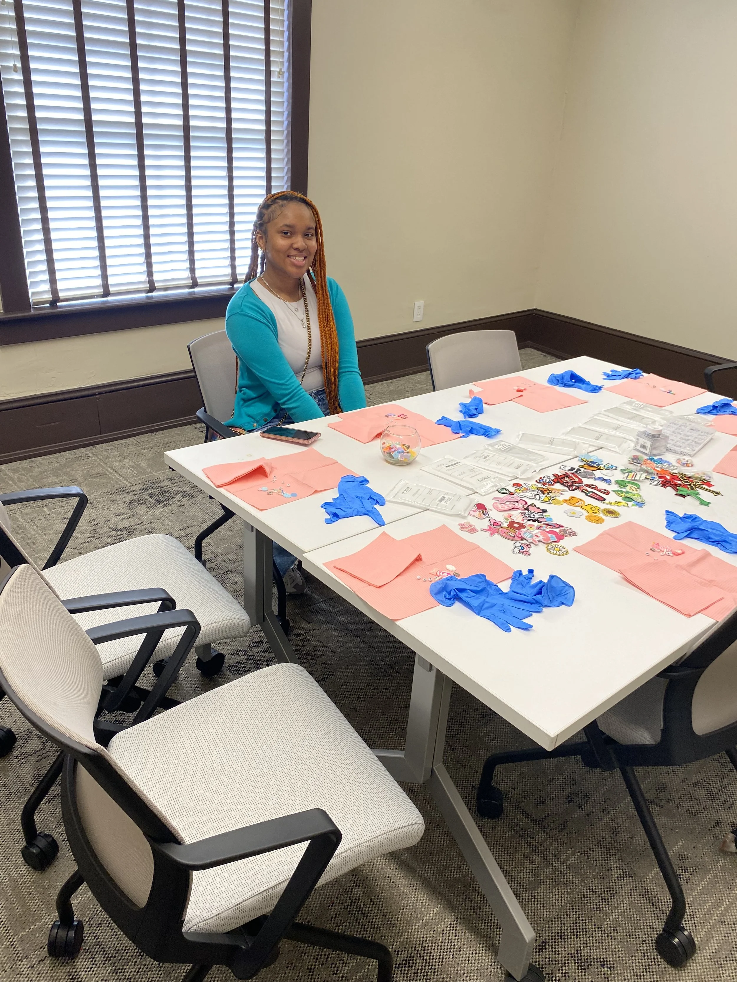 A woman with long reddish braids sitting at a table set up for a craft activity in a conference room, with blue gloves, pink napkins, and various decorations on the table.