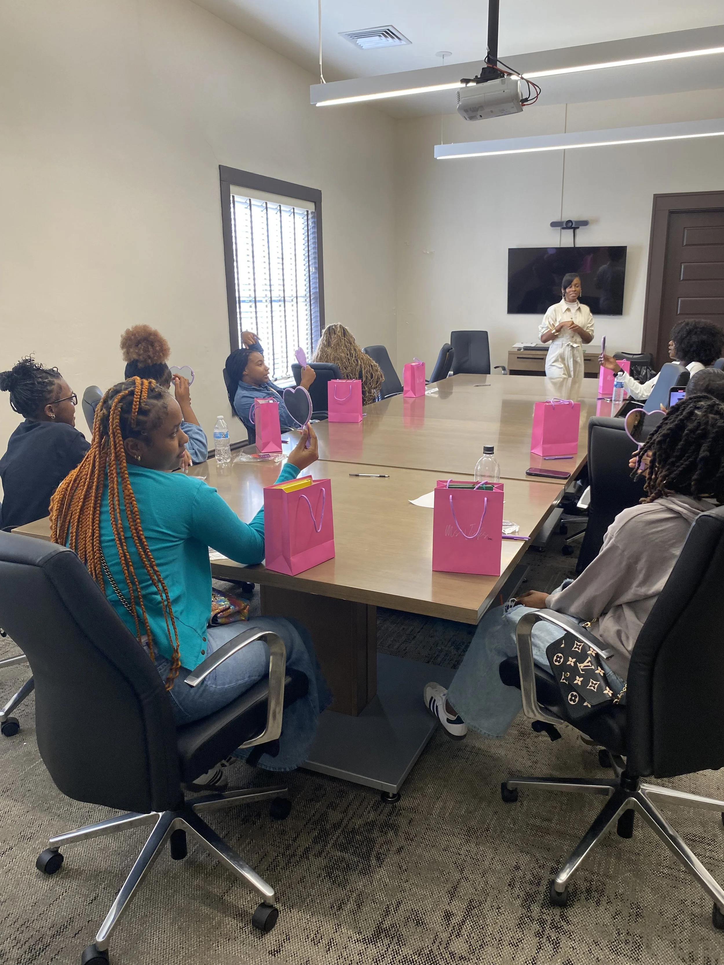 A group of women sitting around a conference table with pink gift bags, listening to a woman standing at the front of the room with a television behind. Some women are taking photos or videos of the speaker.