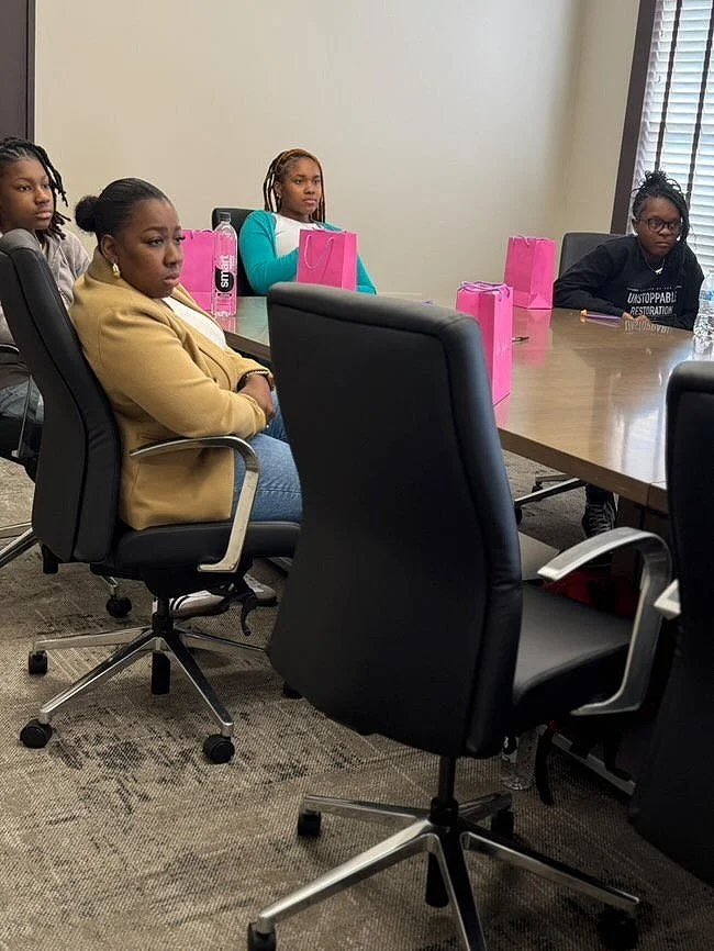Four women sit around a conference table with pink gift bags and water bottles, in a meeting room with window blinds.