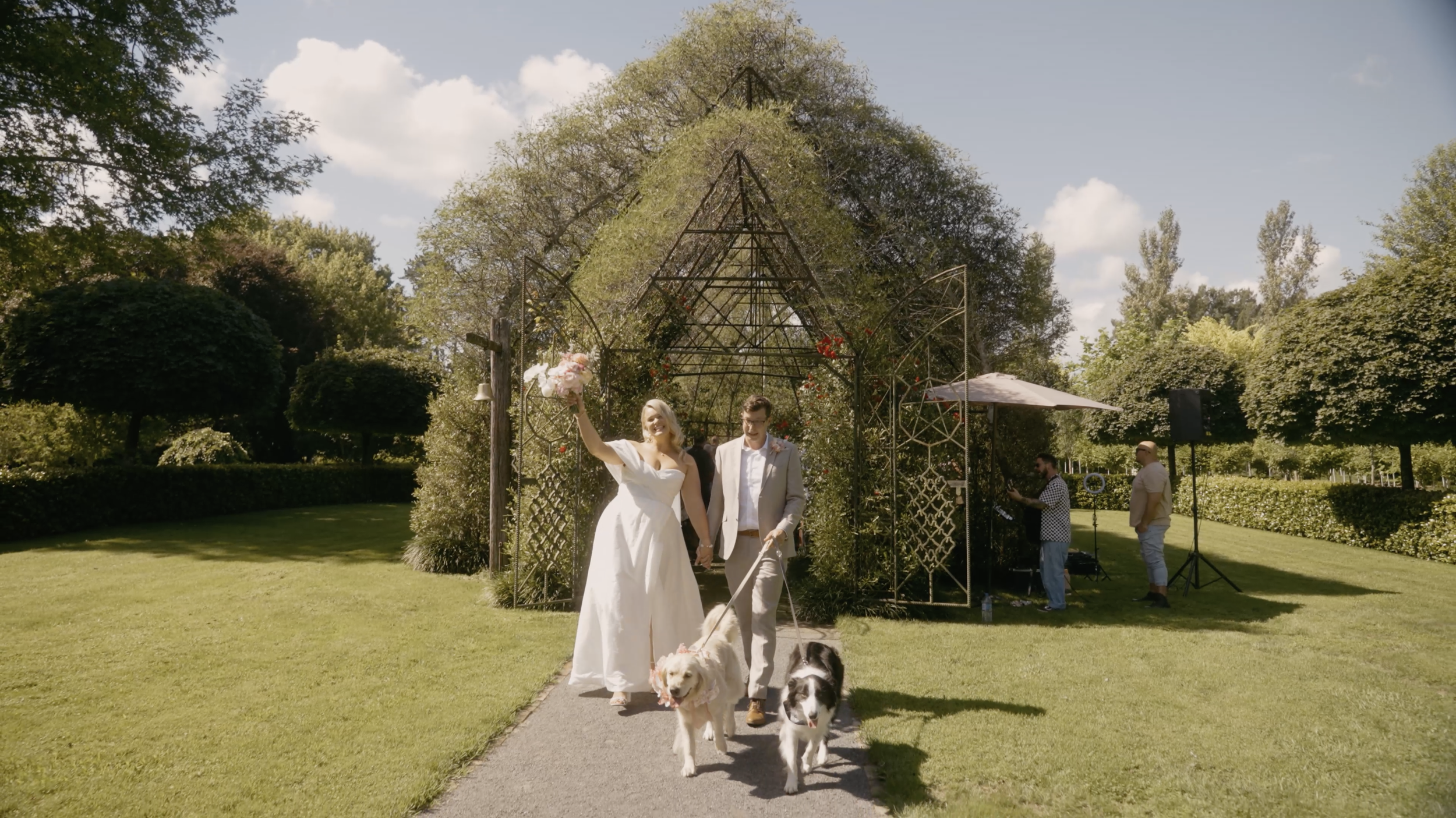 A wedding couple walking outdoors with two dogs, holding hands, in front of a decorative garden arch, greenery, and trees under a partly cloudy sky.
