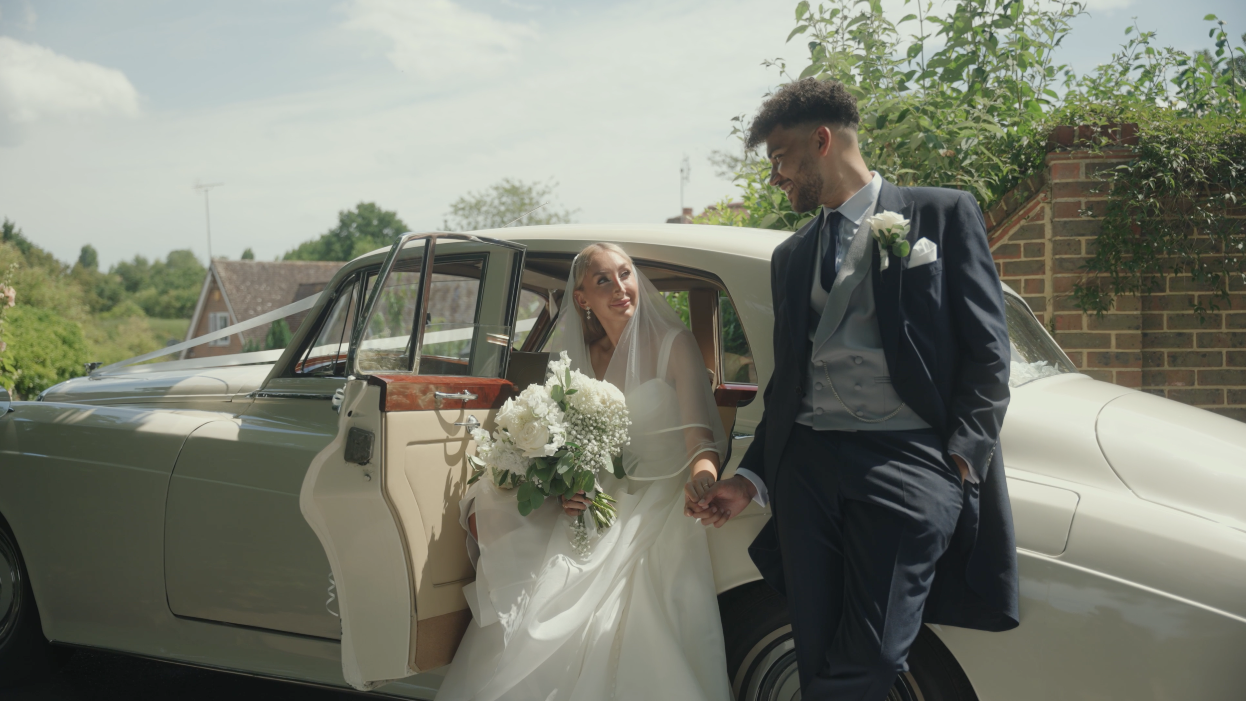 A bride sitting in the backseat of a vintage white car, holding a bouquet of white flowers, and a groom standing outside, holding her hand and looking at her, outdoors on a sunny day.