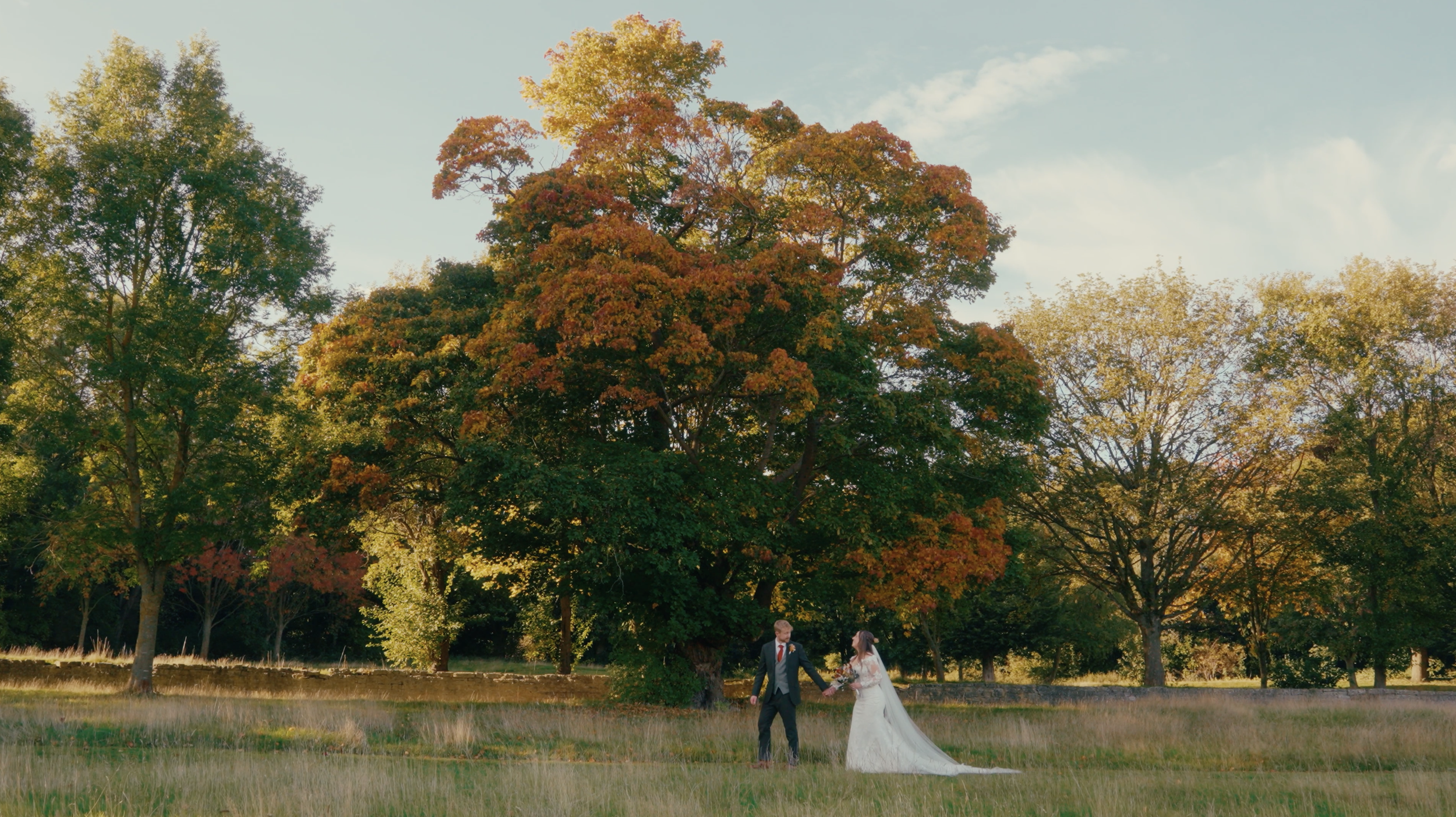 A bride and groom holding hands while standing on a grassy field with trees showing fall foliage in the background.