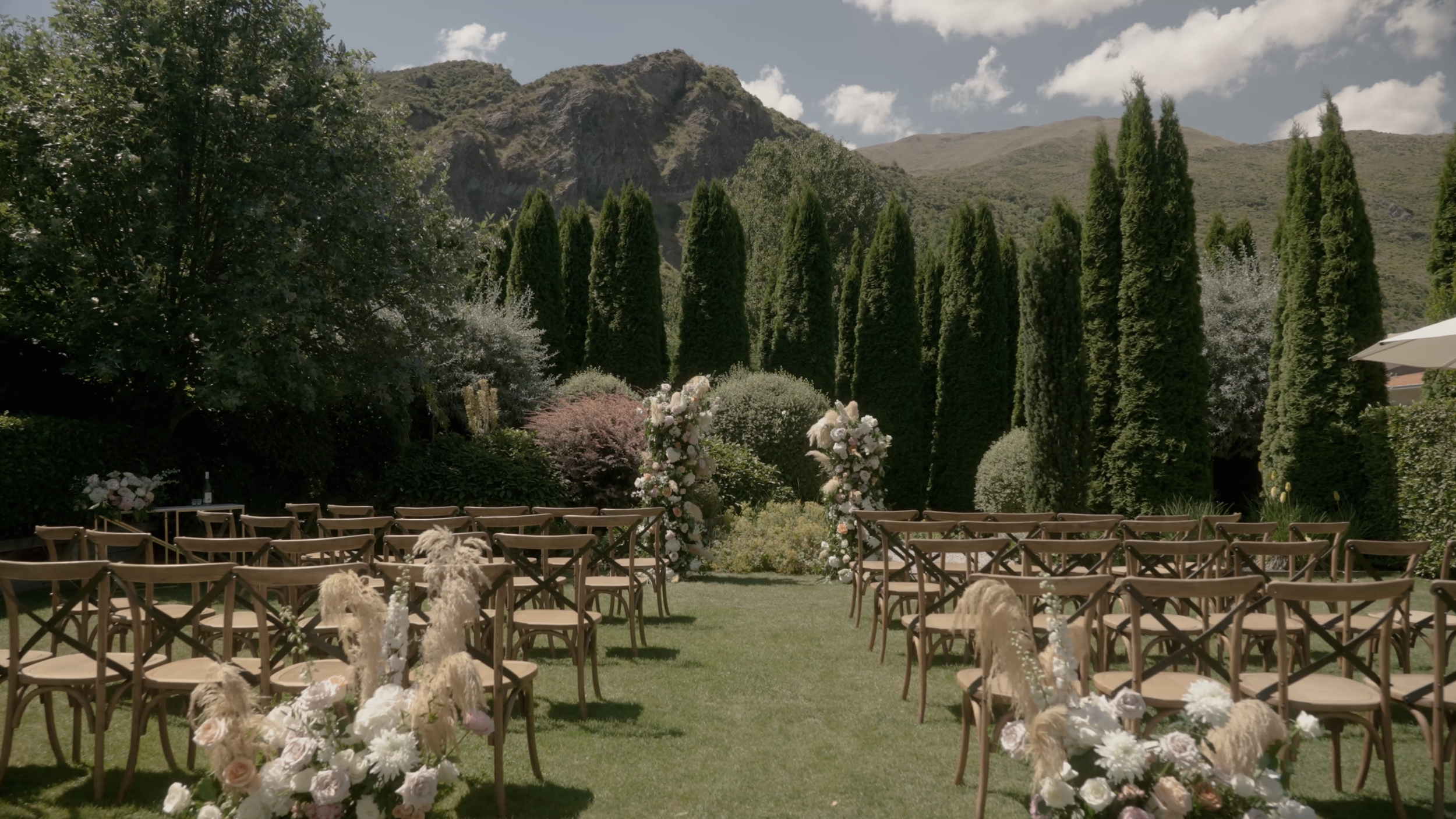 Outdoor wedding ceremony setup with chairs arranged on grass, floral decorations, and a backdrop of tall trees and mountains under a partly cloudy sky.