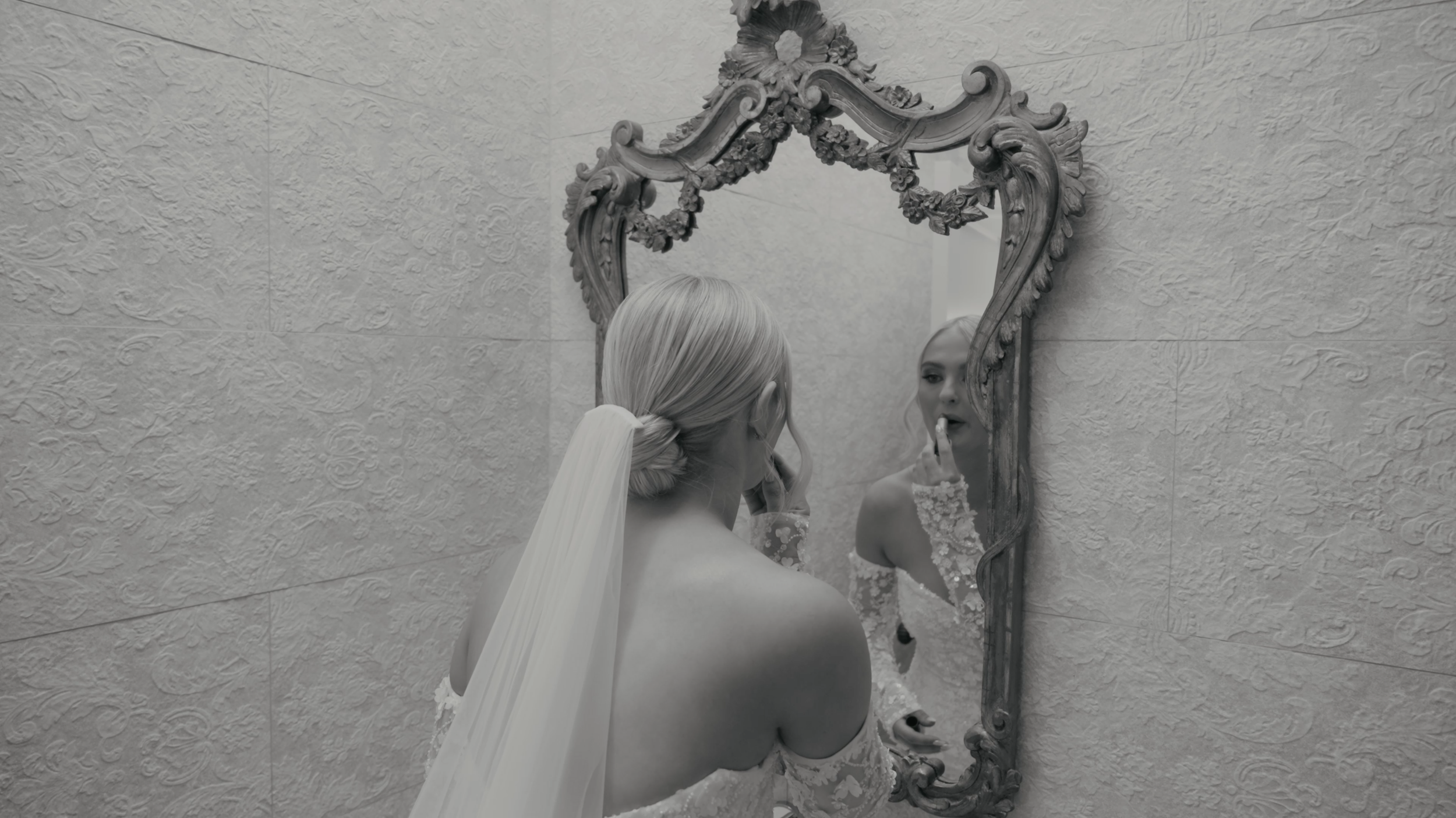A woman in a wedding dress and veil looks at her reflection in an ornate mirror, touching her lip while preparing for her wedding.