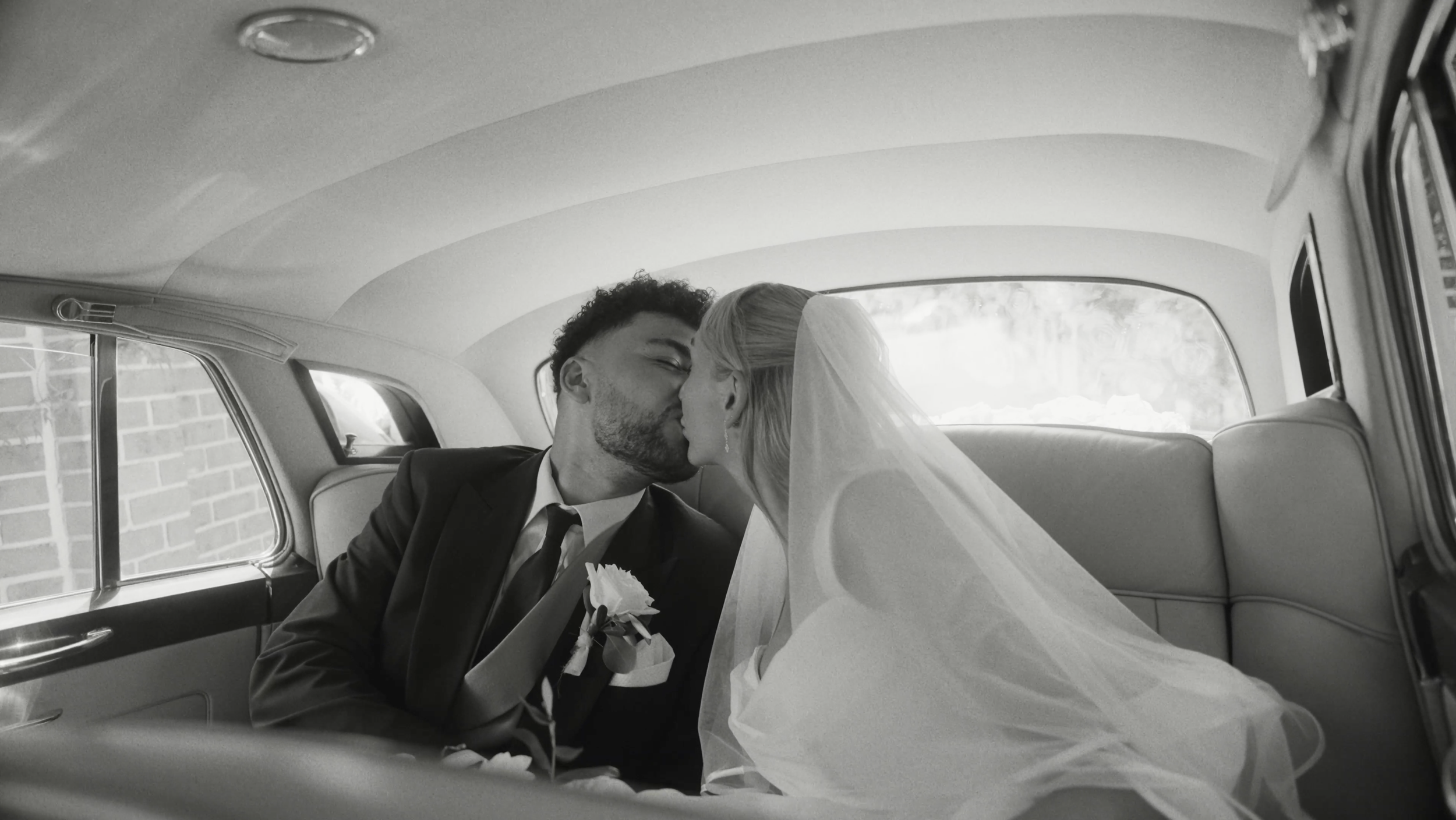 A newlywed couple sharing a kiss inside a classic car, with the bride wearing a veil and the groom in a tuxedo with a boutonnière.
