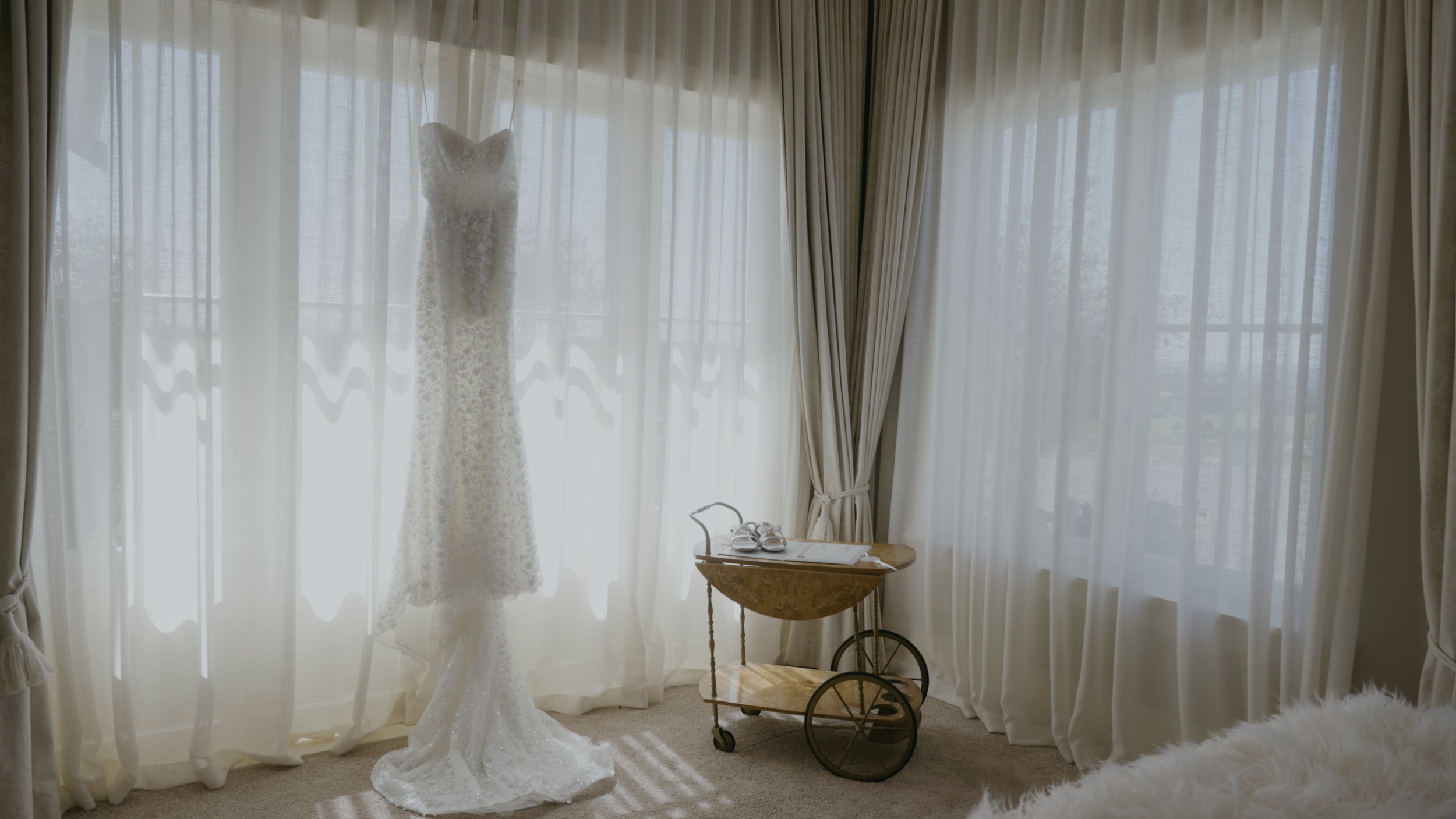 Wedding dress hanging in front of sheer curtains, with a small wooden cart holding a pair of shoes and a book nearby.