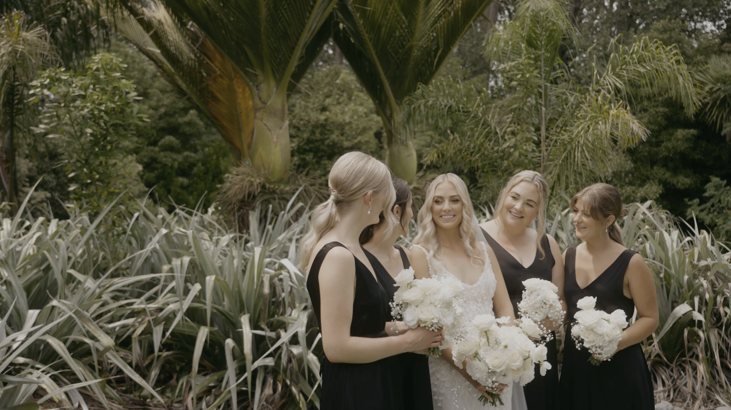 A bride in a white gown holding a bouquet of white roses stands with four bridesmaids in black dresses, holding white floral bouquets, in a lush garden setting.