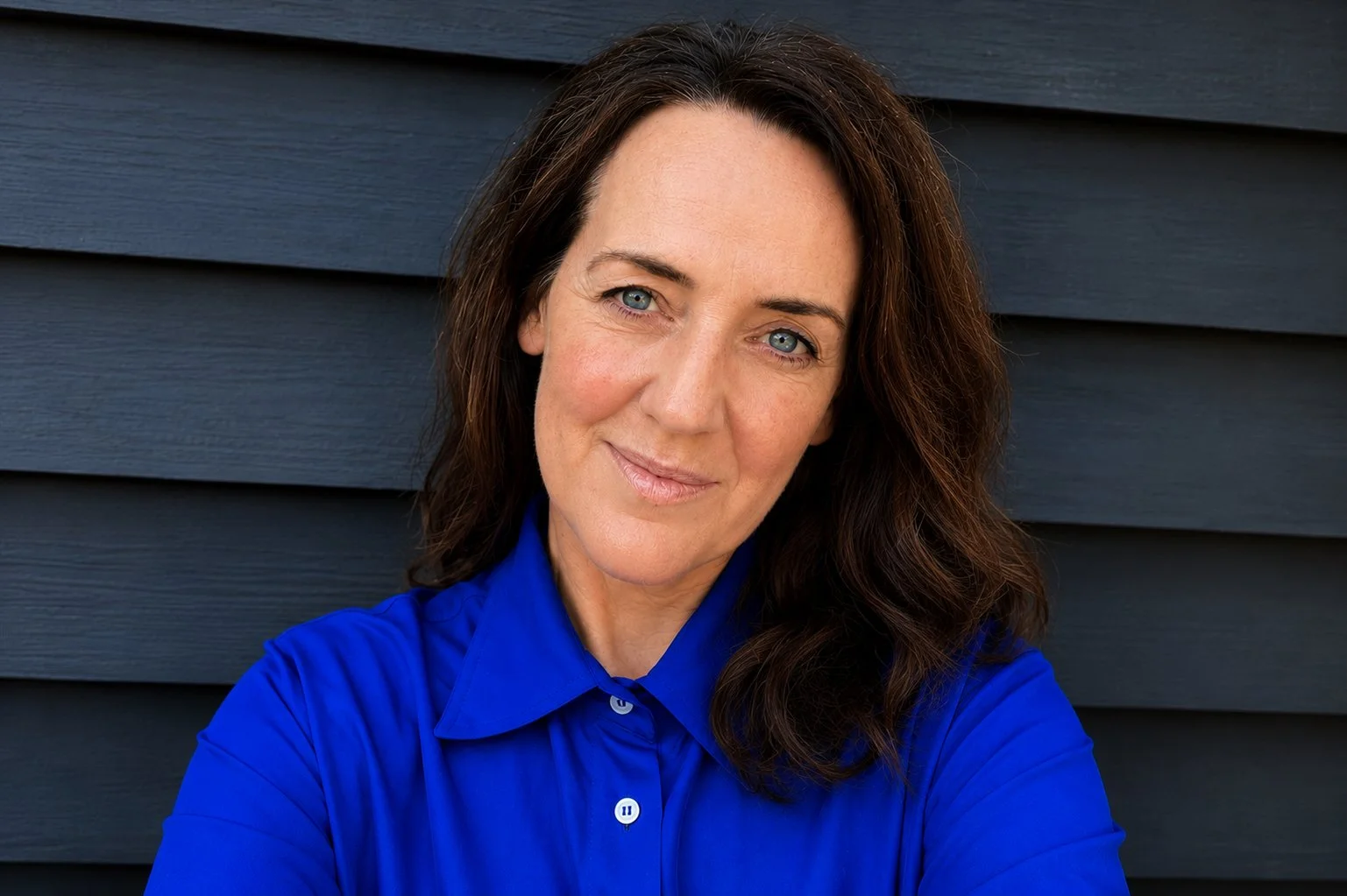 Portrait of a woman with shoulder-length dark brown hair, blue eyes, and light skin, wearing a bright blue button-up shirt, standing in front of a dark gray wooden wall.