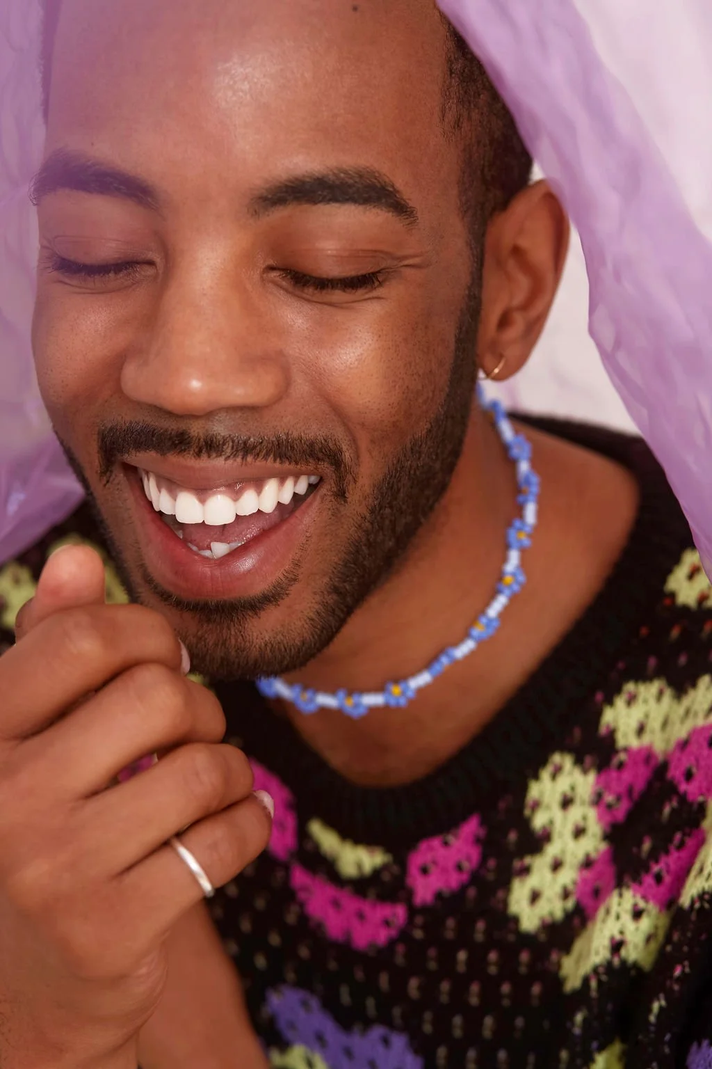 black man smiling during portrait photo session in colourful sweater and purple flower necklace under purple fabric