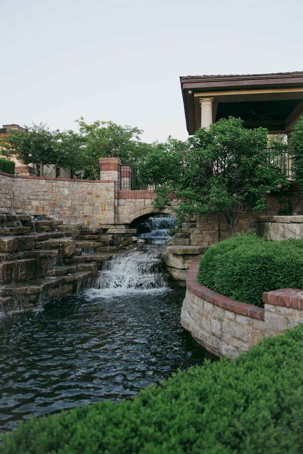 Stone waterfall with greenery and a small bridge in a landscaped garden.
