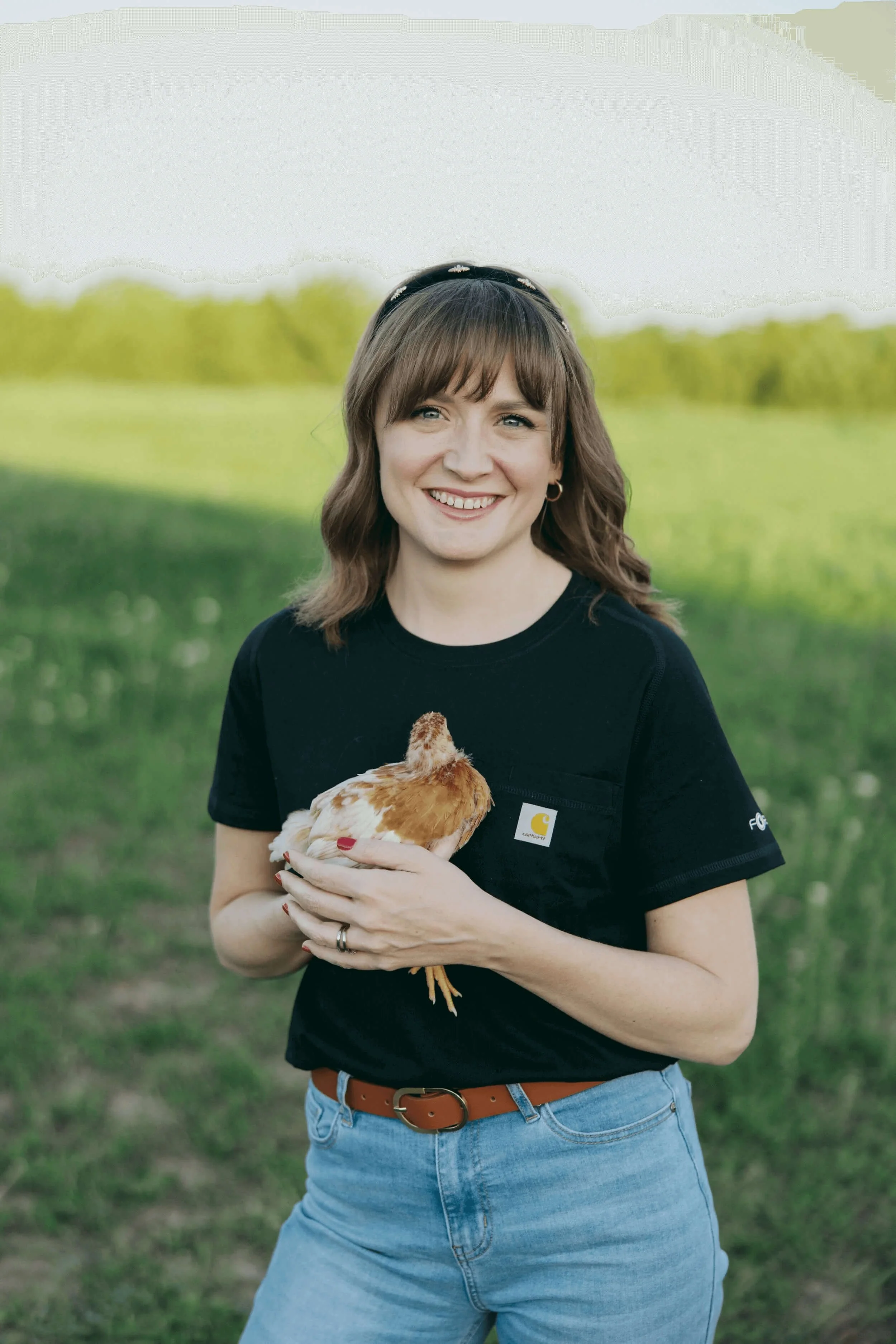 A woman standing outdoors in a grassy field, smiling, holding a chicken with a reddish-brown and white feather pattern.