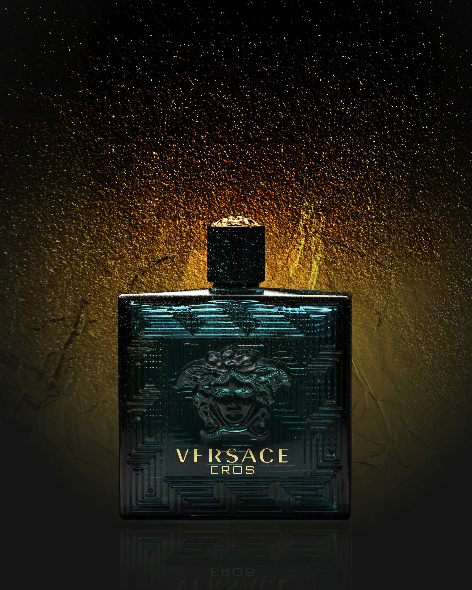 A bottle of Versace Eros perfume with a textured, dark turquoise design and Medusa head emblem on the front, set against a dark, textured background with golden and orange lighting effects.