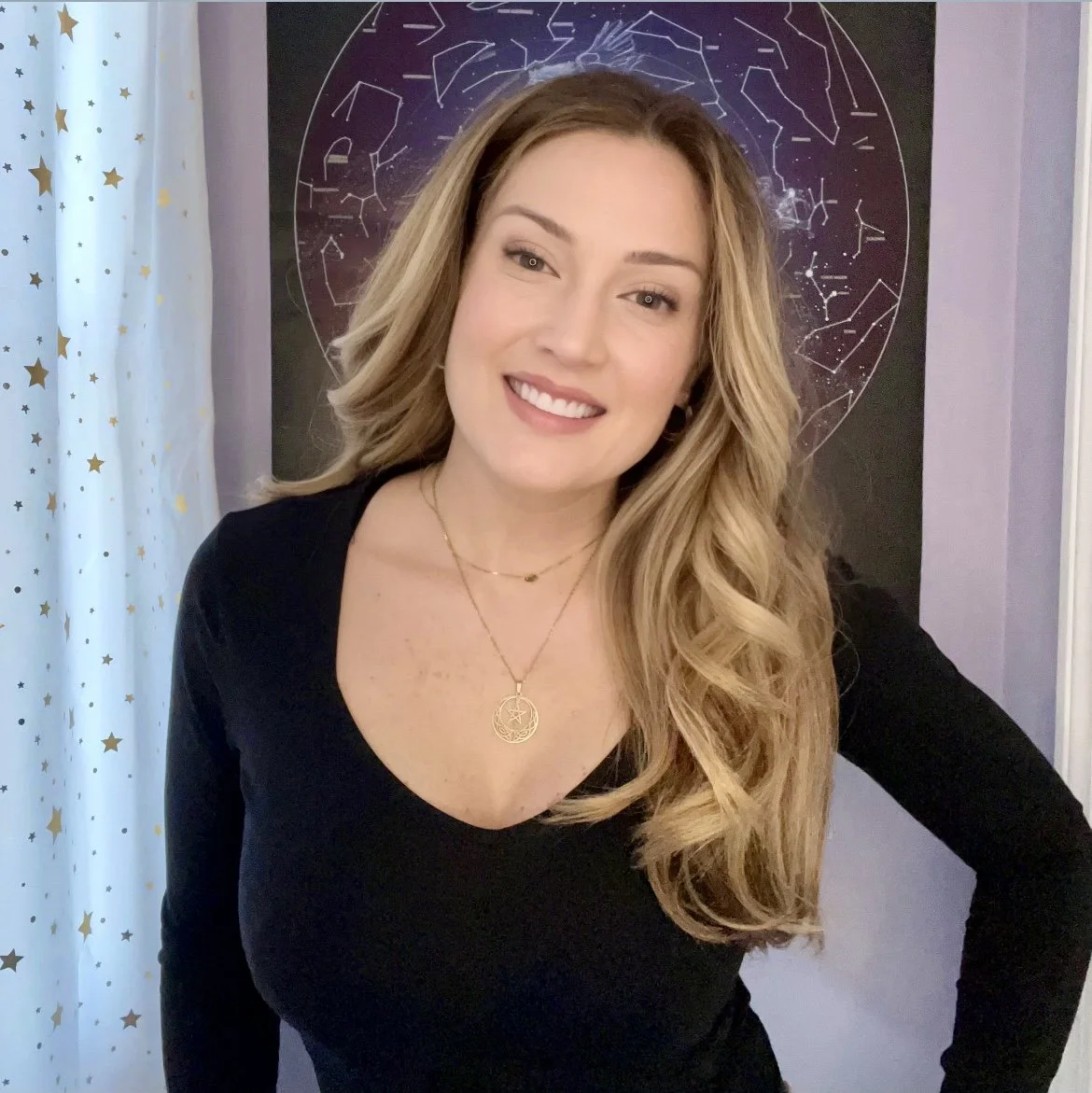 A woman with long blonde hair smiling, wearing a black top and layered necklaces, standing in front of a celestial-themed star chart poster.