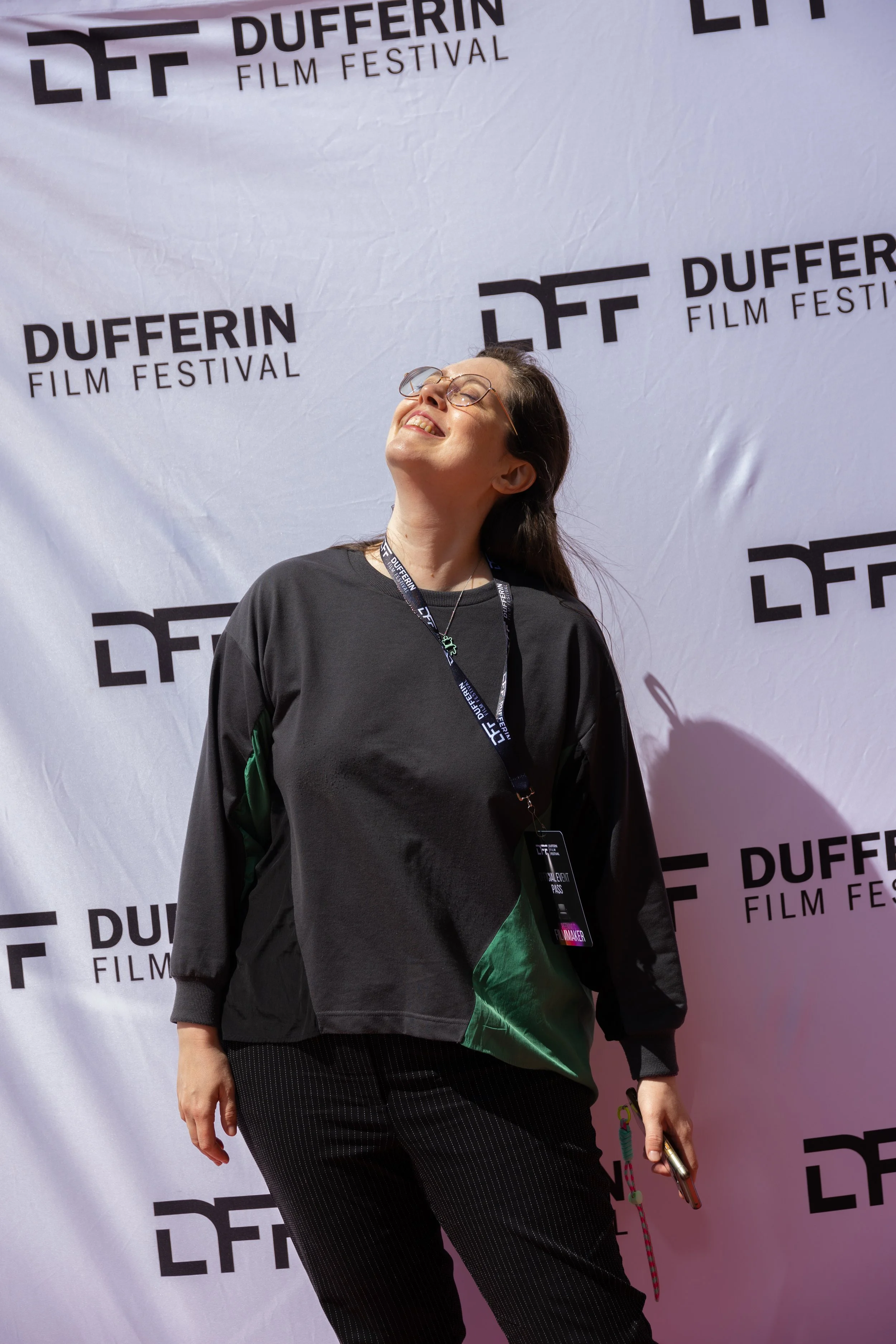 Person standing in front of Dufferin Film Festival backdrop, smiling and looking upwards, holding a phone and wearing a lanyard.