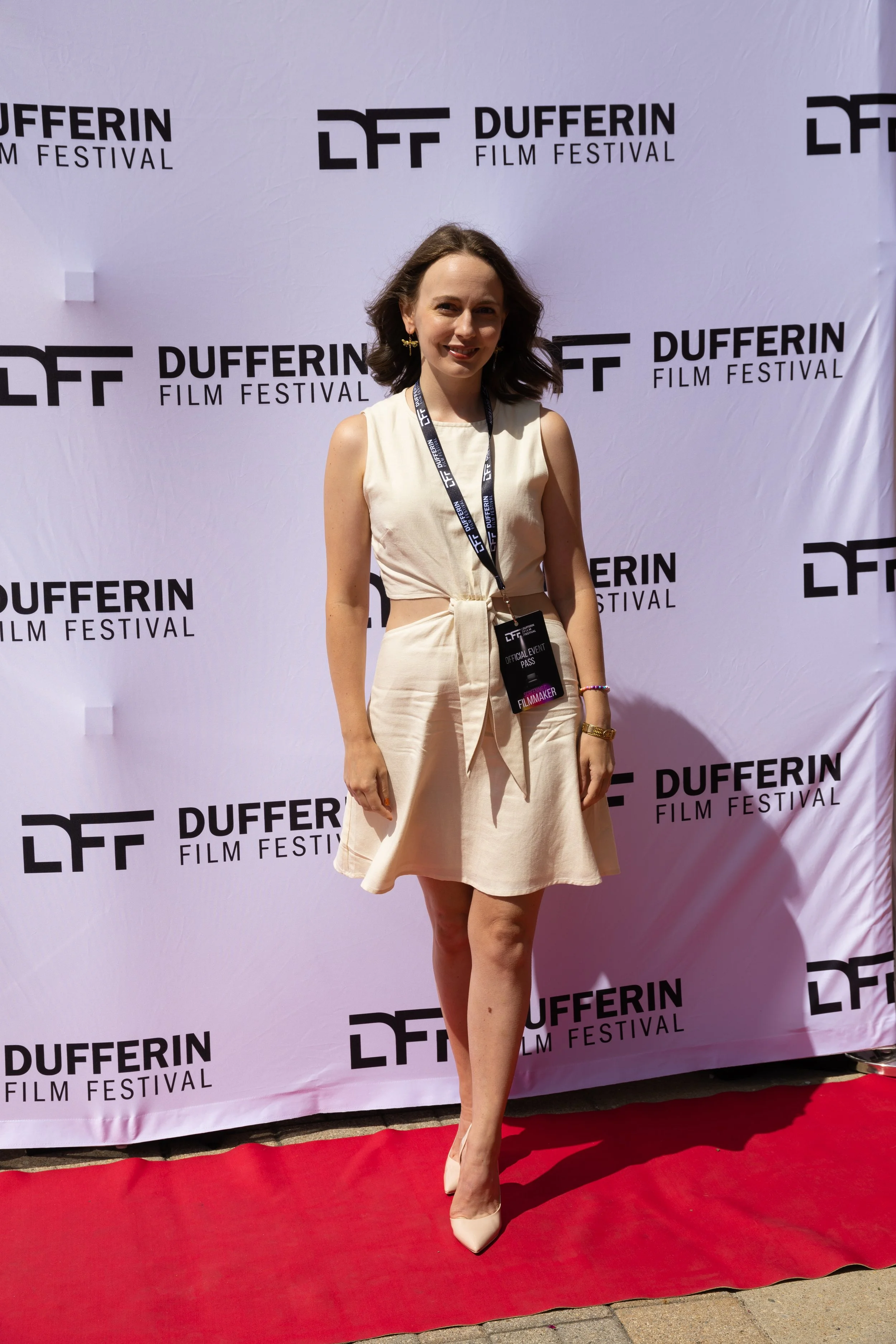 Woman standing on red carpet with Dufferin Film Festival backdrop, wearing a light-colored dress and event pass.