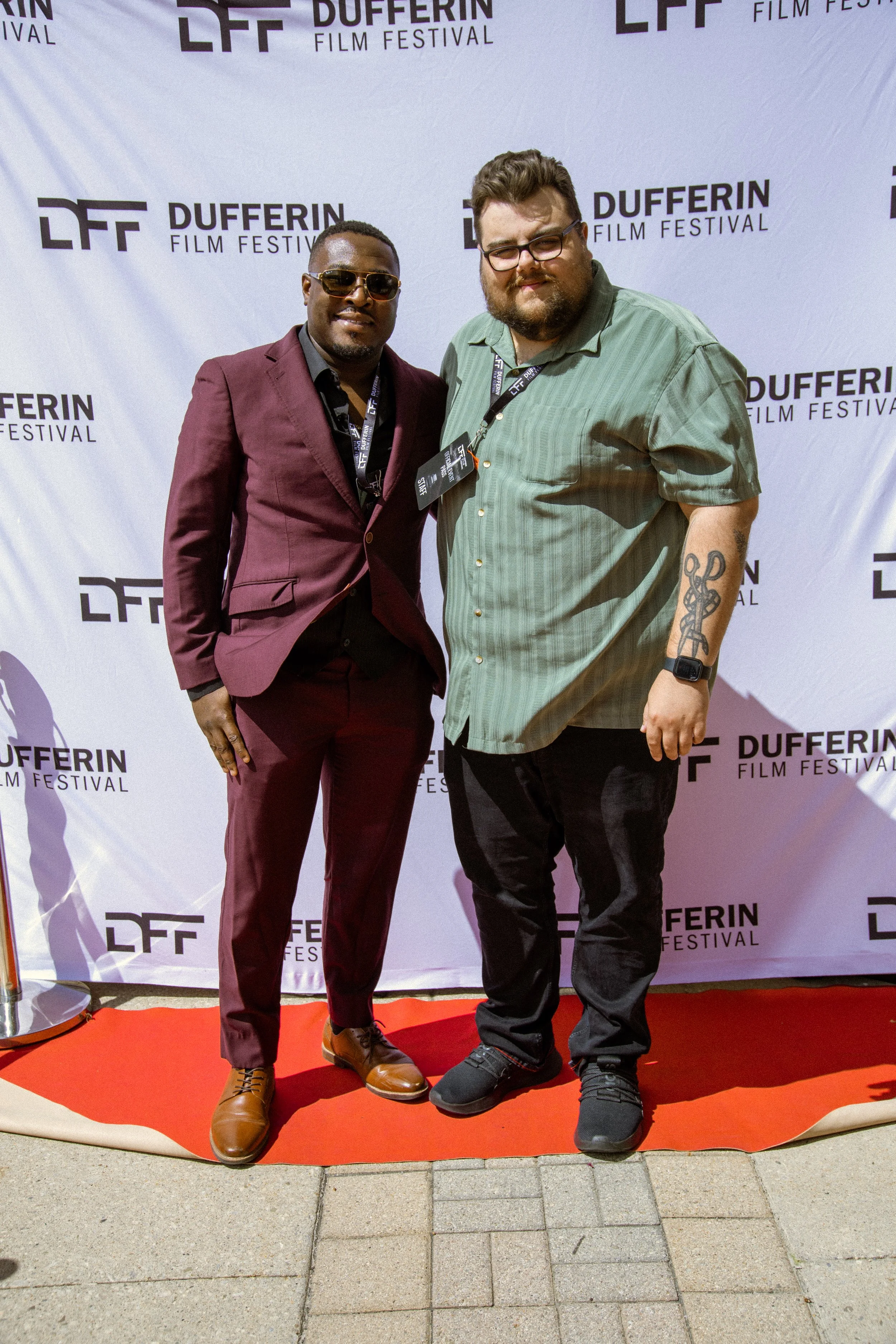 Two men posing on a red carpet at the Dufferin Film Festival. One is wearing a burgundy suit and sunglasses; the other is in a green shirt with glasses.
