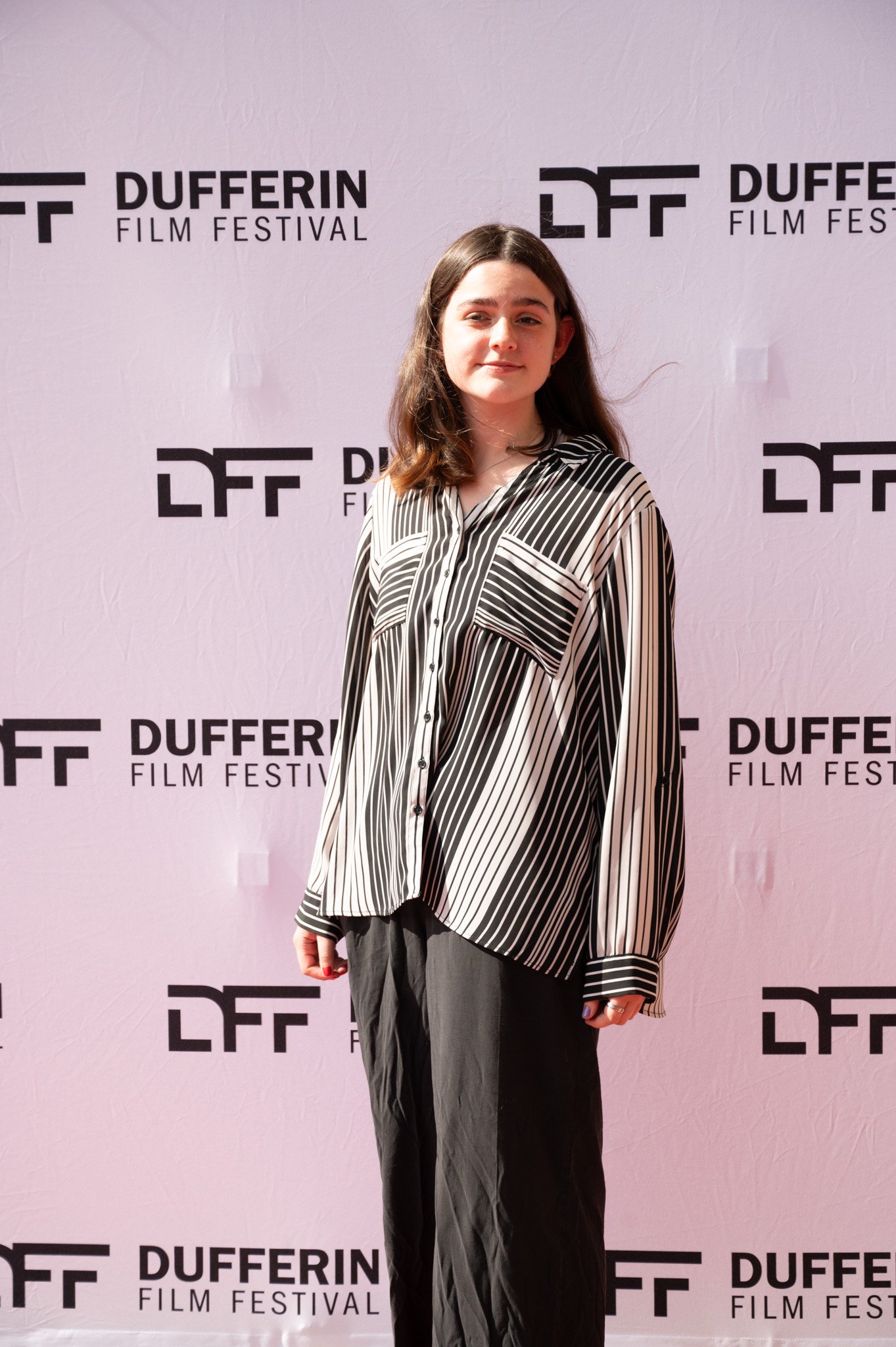 Person at Dufferin Film Festival standing in front of a branded background, wearing a black and white striped shirt and black pants.