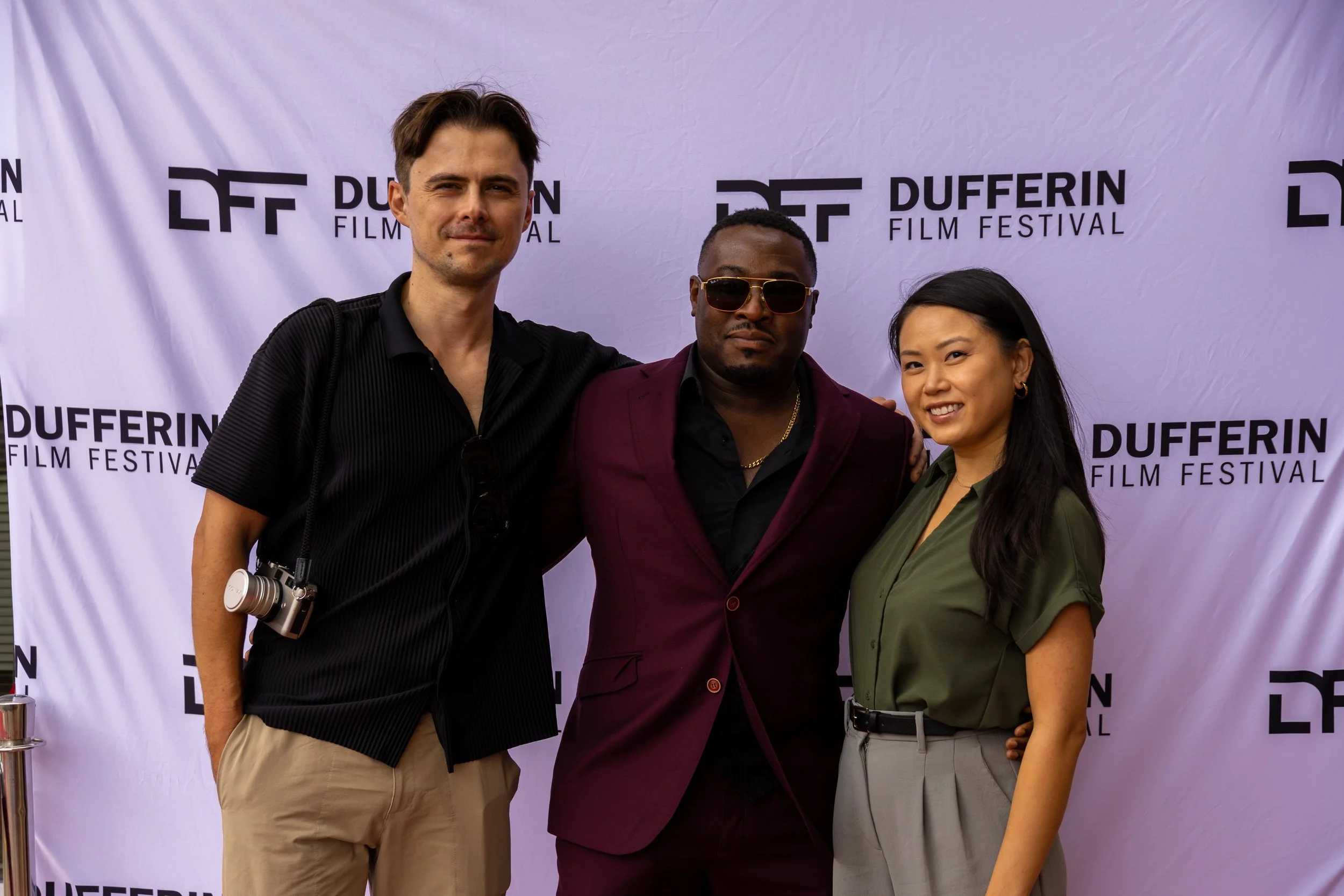 Three people posing in front of a Dufferin Film Festival backdrop; two men and a woman, one man holding a camera and wearing sunglasses, the woman in a green blouse and gray pants.