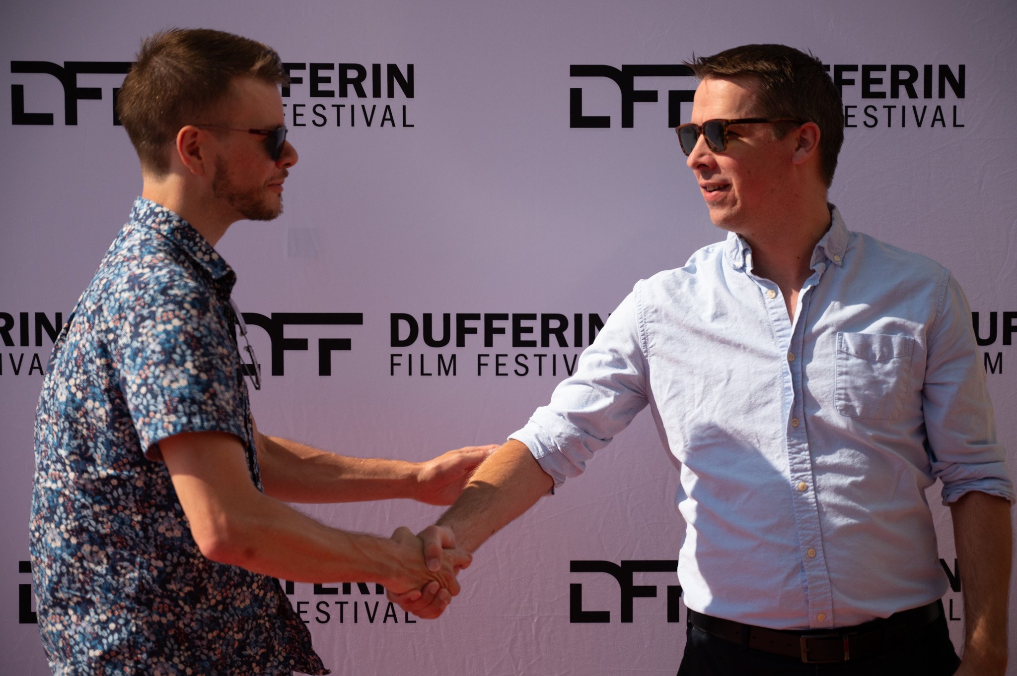 Two men shaking hands at the Dufferin Film Festival, both wearing sunglasses. One man is in a blue floral shirt, and the other in a light blue button-up shirt. They are standing in front of a festival step and repeat banner.