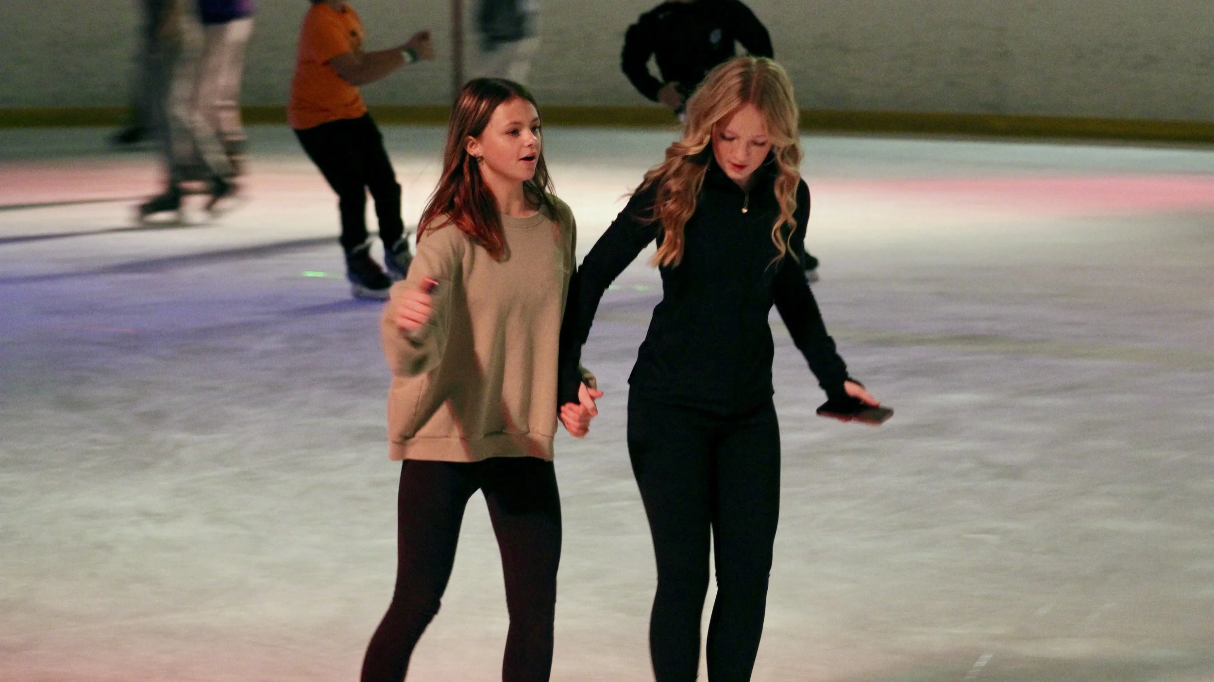 People ice skating at an indoor rink, holding hands, with others skating in the background.