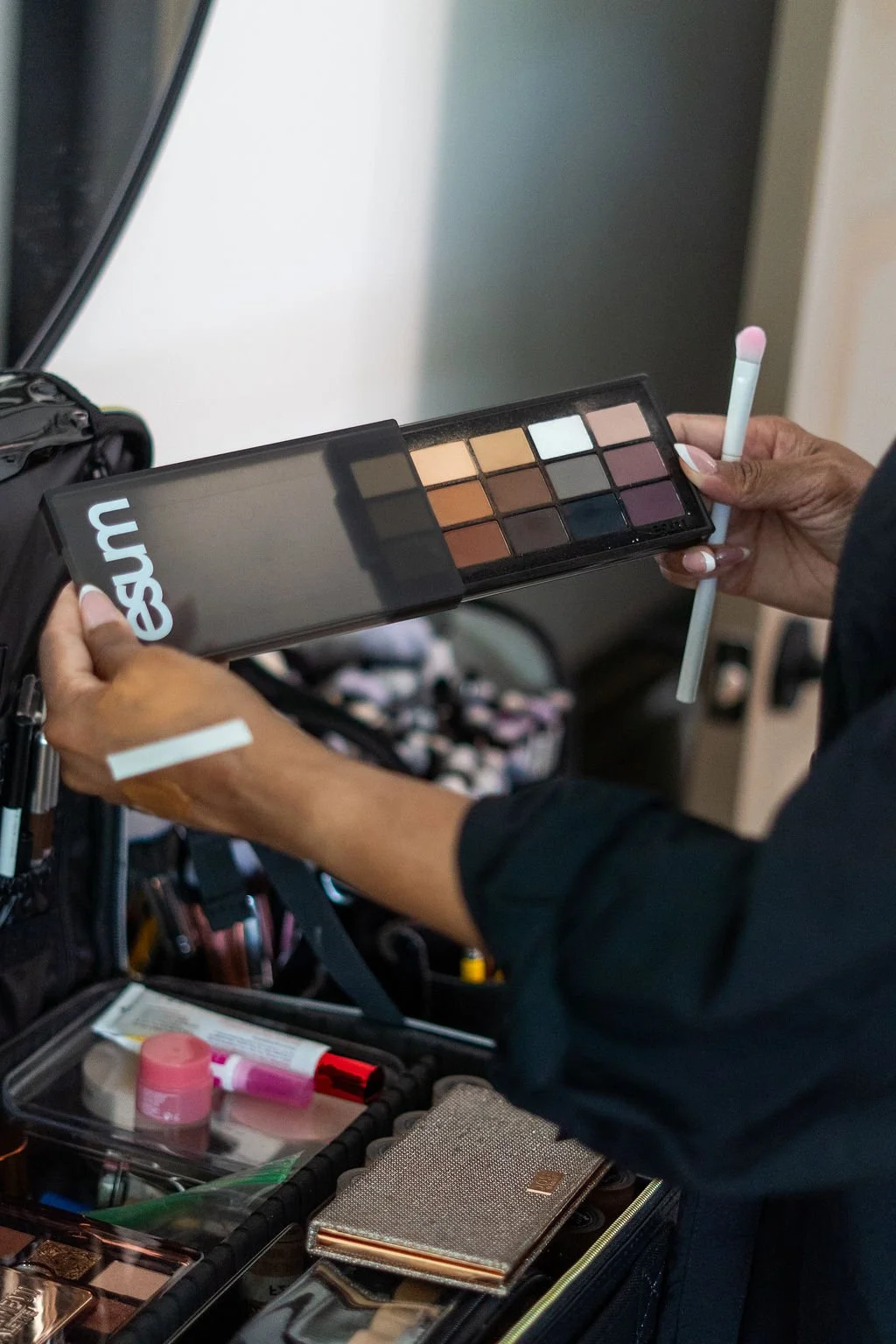 Person holding an eyeshadow palette with multiple shades and a makeup applicator, with a makeup bag and cosmetic items on a table.