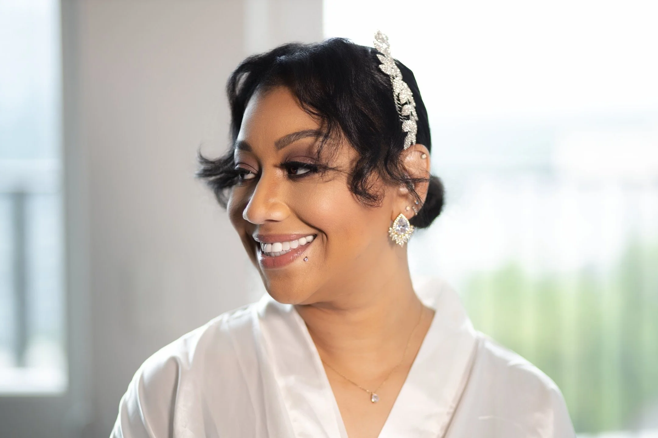 A woman with short black hair, styled in waves, wearing large sparkling earrings, a diamond hair ornament, and a white top, smiling softly in a softly lit room.
