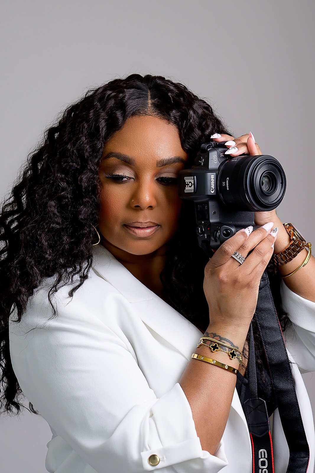 A woman with curly hair holding a Canon camera, wearing a white blazer and gold jewelry, against a plain gray background.