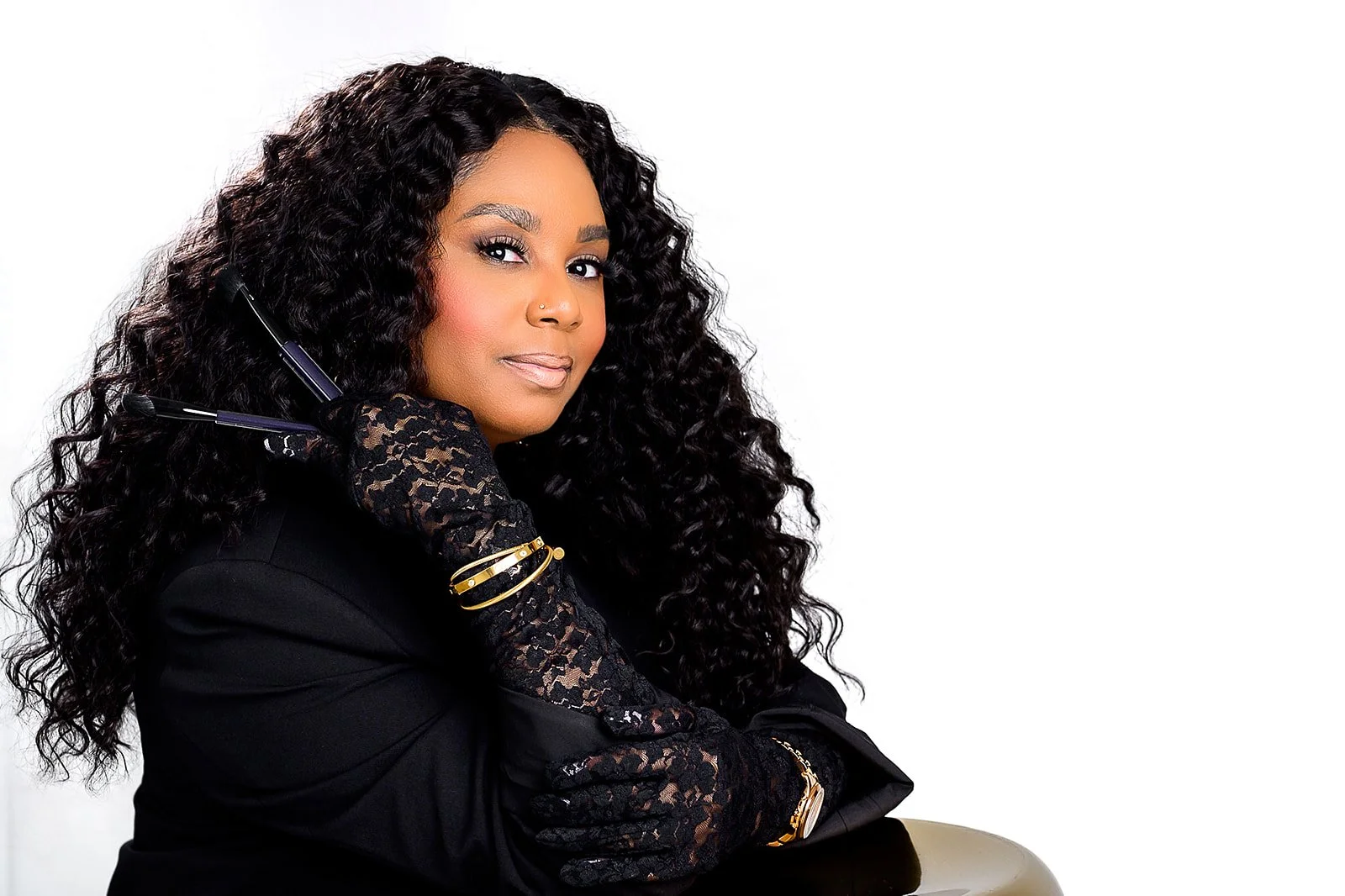 A woman with long, curly black hair wearing a black outfit with lace sleeves, gold jewelry, and black gloves, holding a black telephone receiver to her ear against a plain white background.