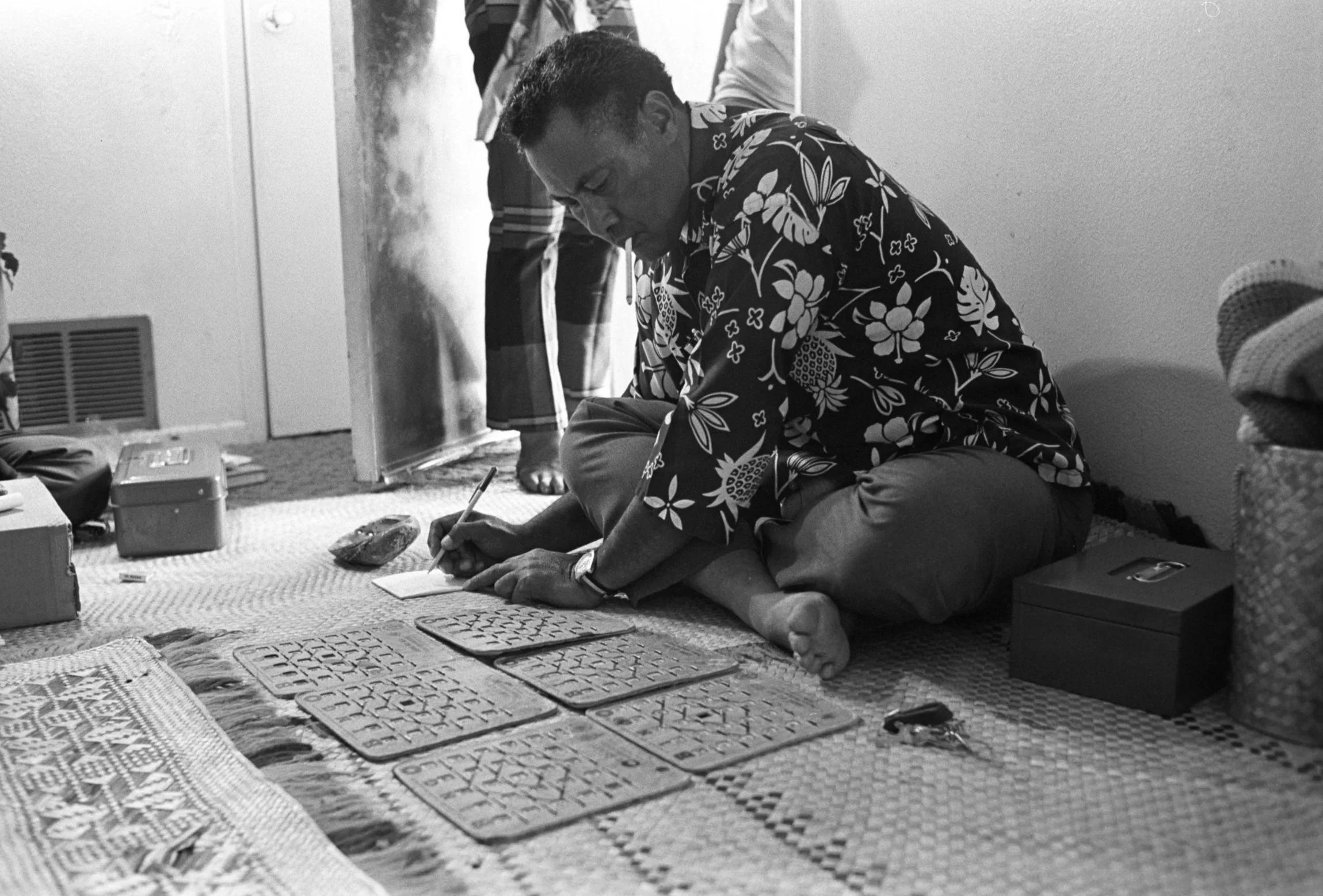 Faces (1972) – Sialoi Residence,
An individual wearing a floral shirt sitting on the floor tallying up the results into a notebook for a bingo game.
Photographed by Alan Ohashi
