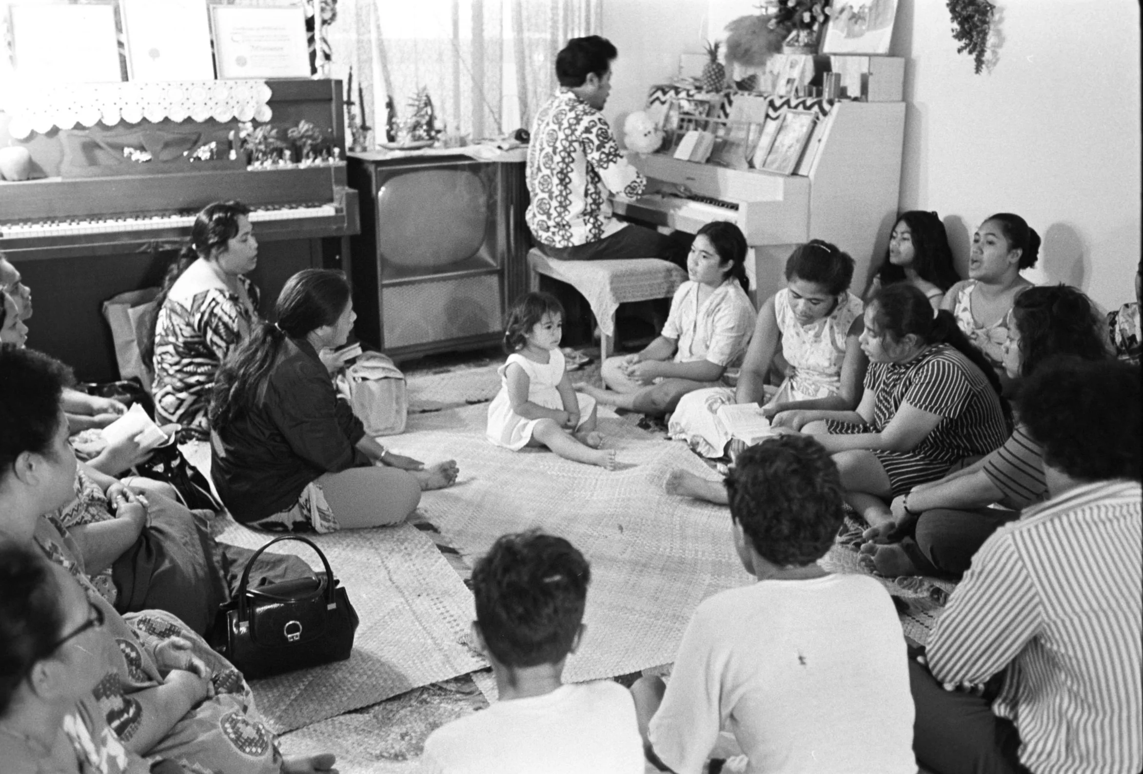 Congregation Sitting on Floor in Reverend (1972) – Sialoi Residence, 
(Samoan Church Service) Men, women, and children of the congregation sitting on the floor during a group practice in the residence of Reverend Sialoi, accompanied by a man playing 