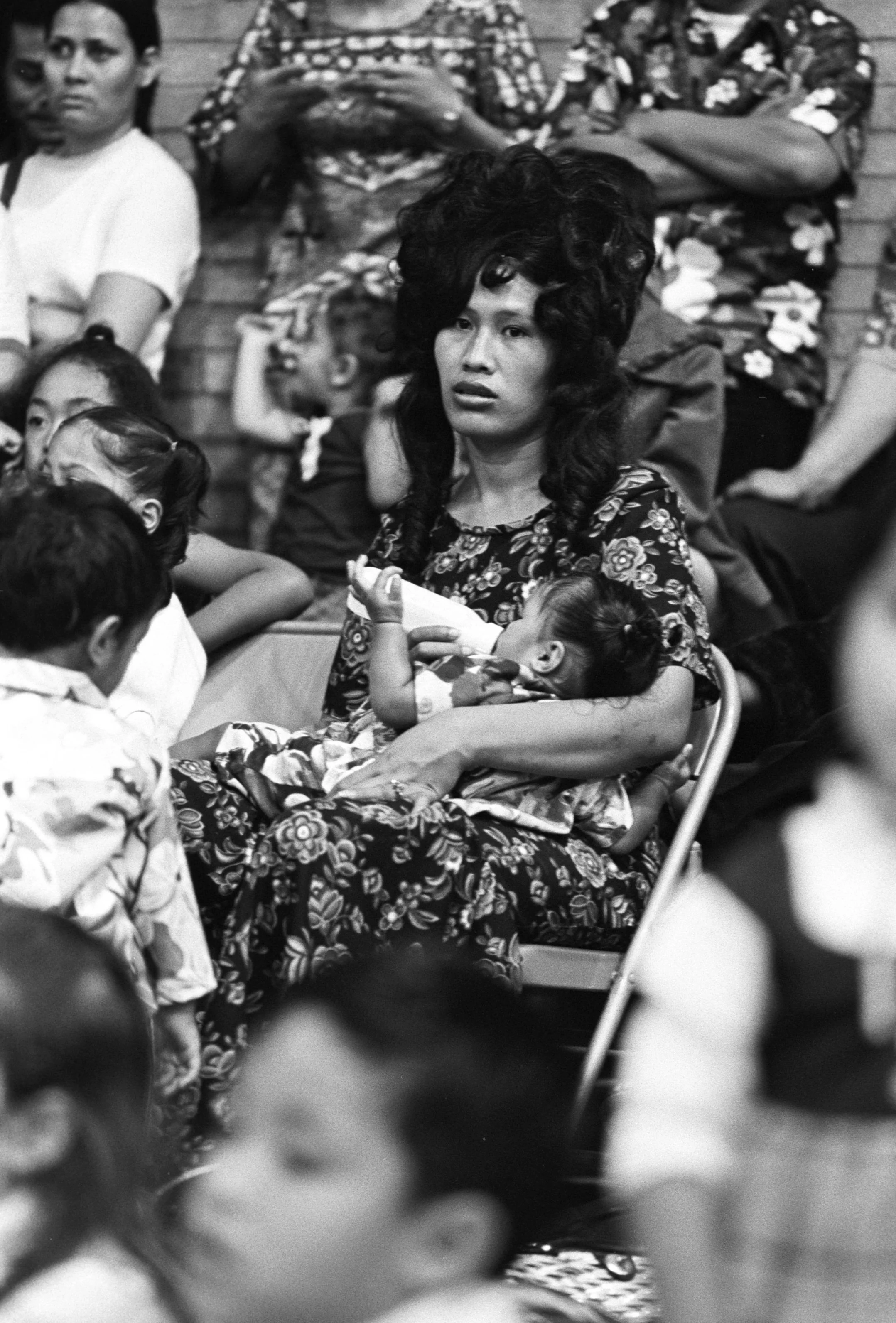 Luau Audience Watching Dancers (1972),
A seated woman feeding an infant among other audience members, watching a traditional dance performance at a luau.
Photographed by Alan Ohashi