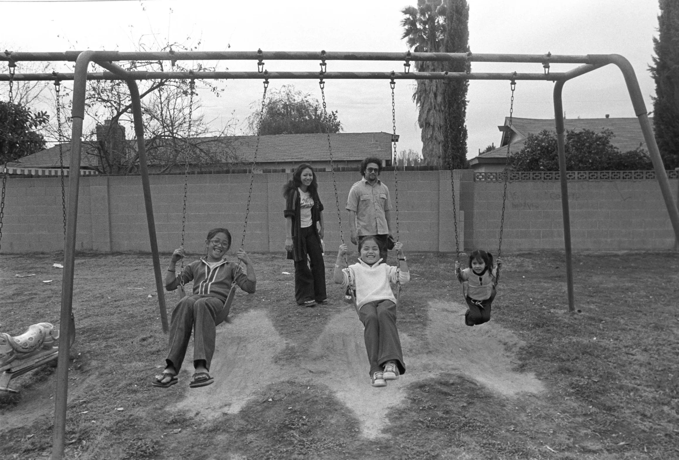 Adults and Children at an Outdoor Swingset (1972)
Two adults standing and three children swinging at a swingset outside.
Photographed by Visual Communications